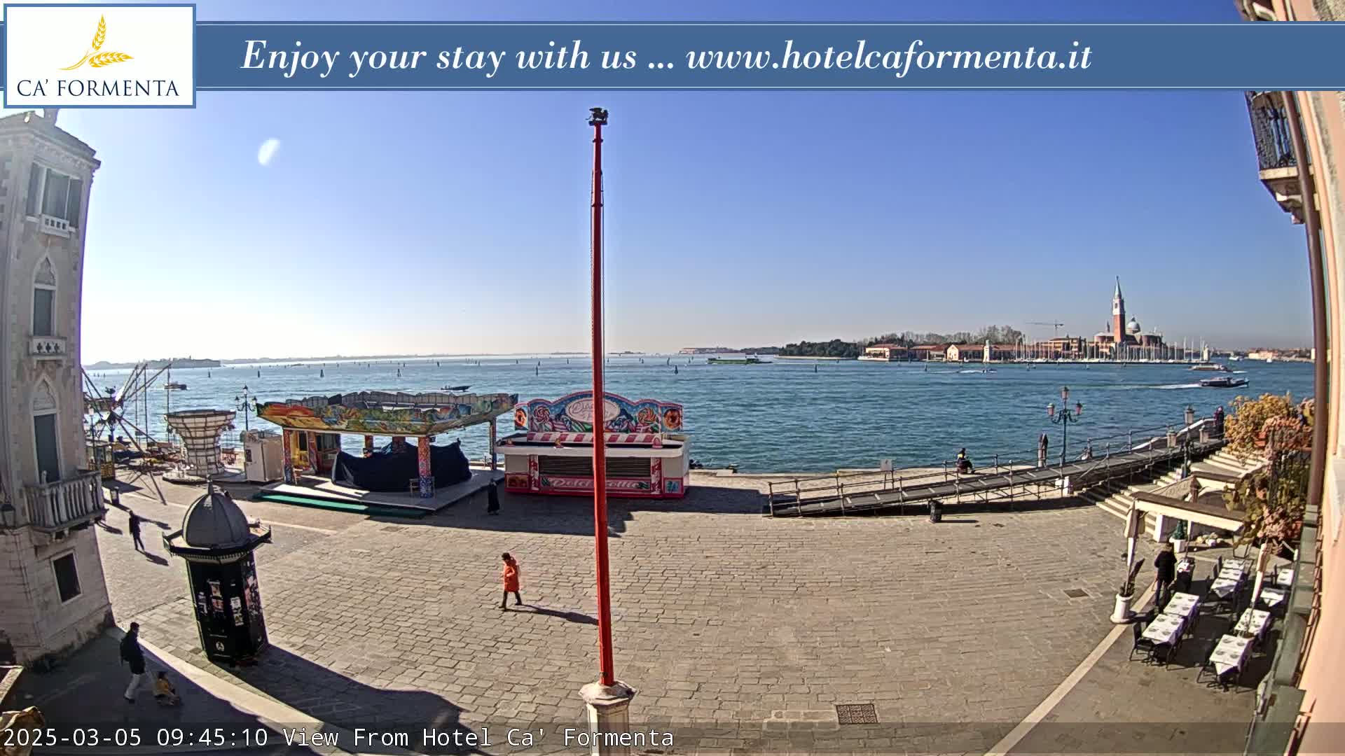 A sunny outdoor scene shows a paved plaza with a small amusement park, a waterside walkway, and a distant view of a city across a calm body of water.