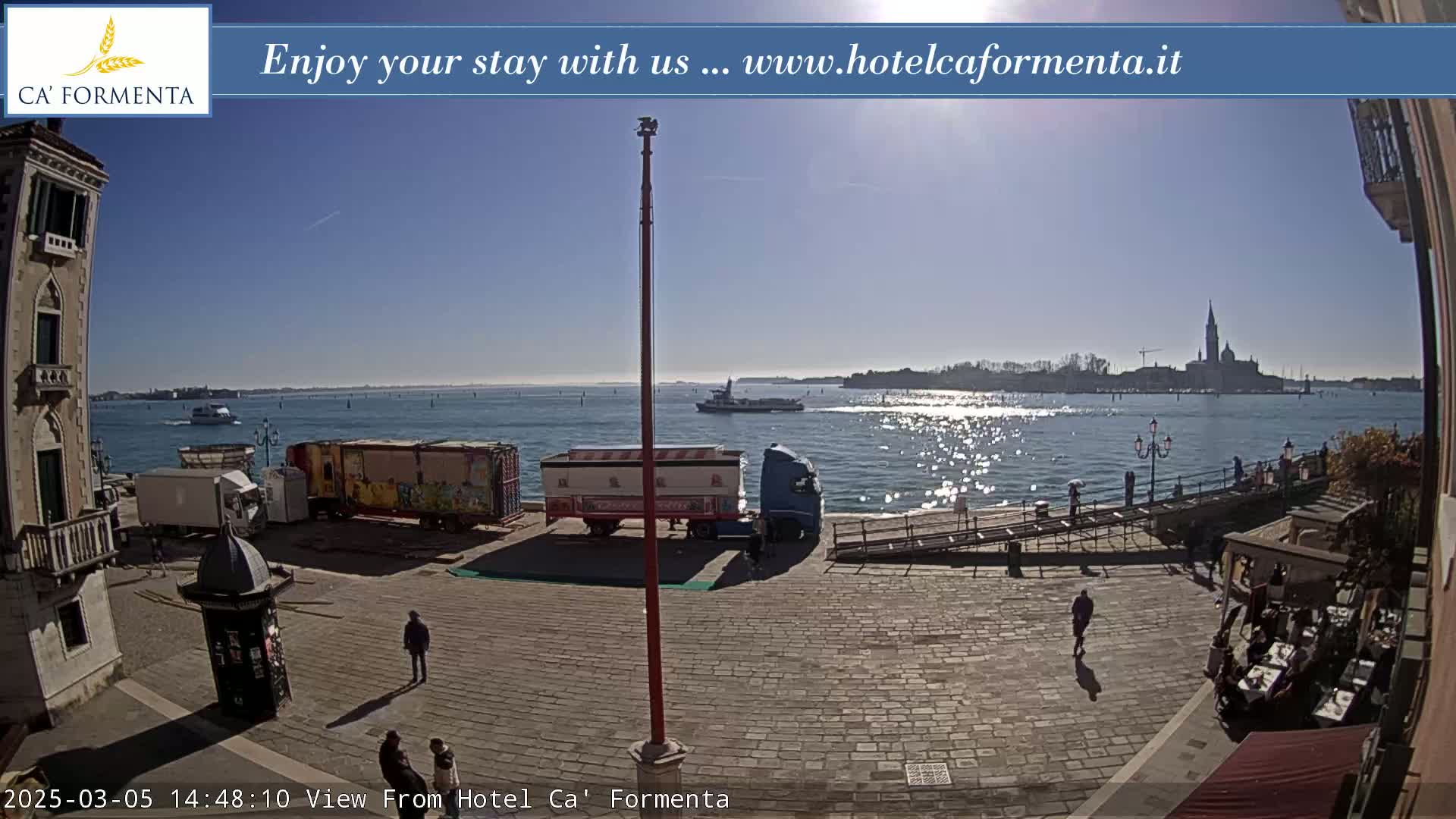 A sunny day reveals a waterfront paved plaza with several people and trucks, overlooking a body of water where a boat is visible, and an island with buildings in the distance.