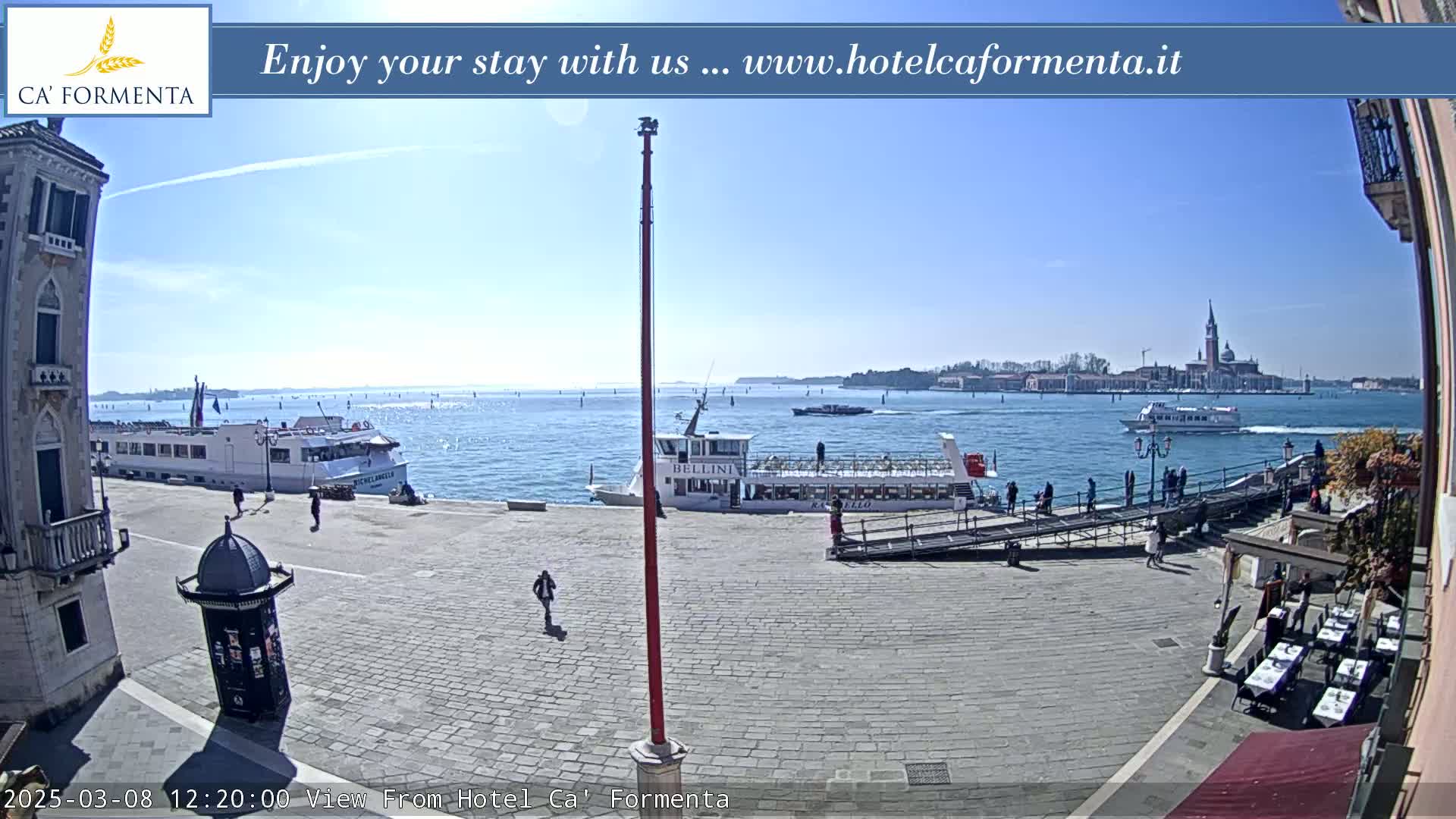 A sunny Venetian waterfront scene shows several boats docked near a plaza with people walking and outdoor seating.