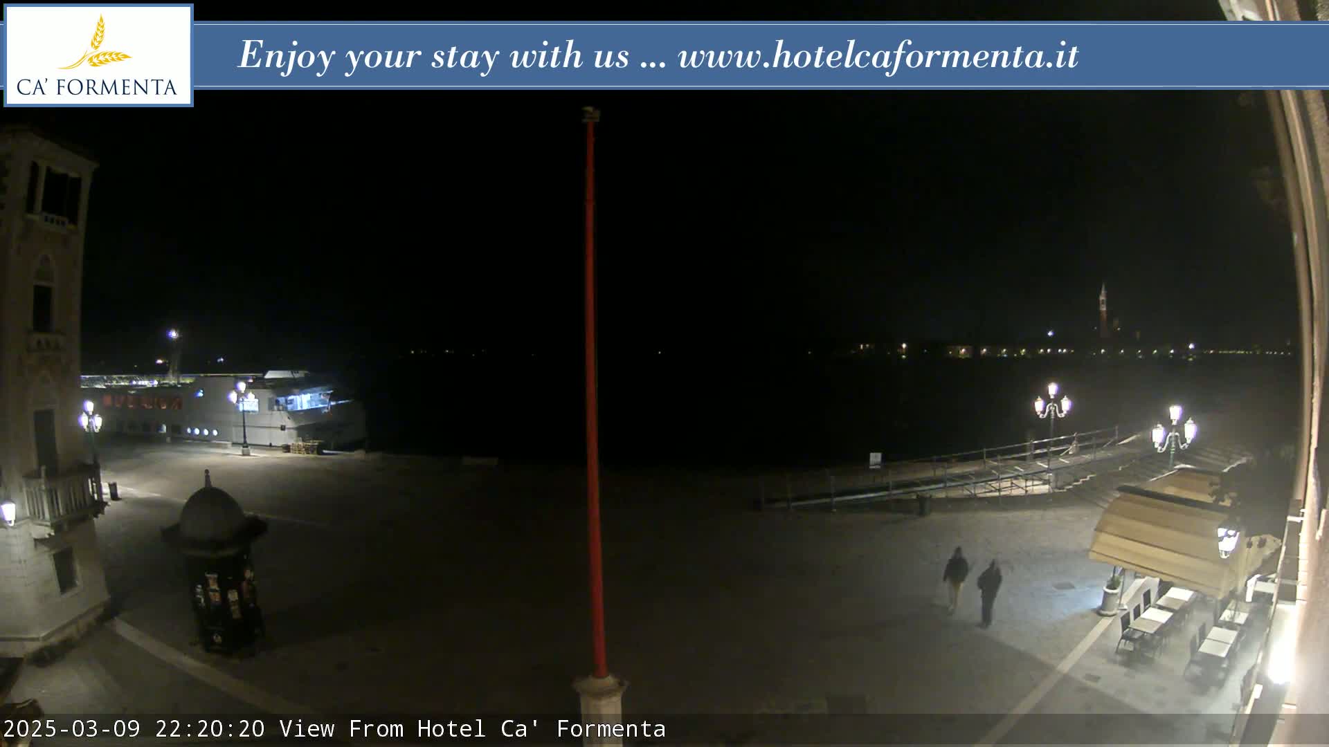 A nighttime view of a waterfront plaza with a boat docked, two pedestrians, and outdoor seating at a restaurant, under a clear, dark sky.