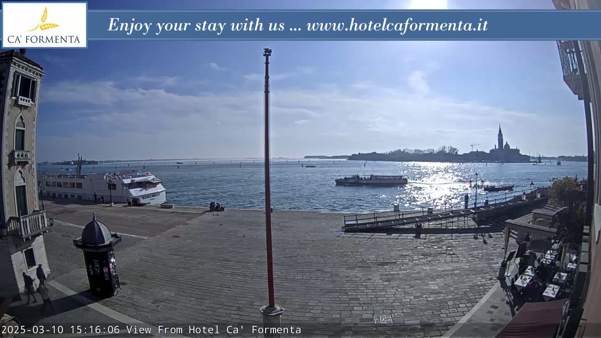 A sunny day shows a paved waterfront area with a large boat docked, smaller boats in the water, and a distant view of a city across the water.