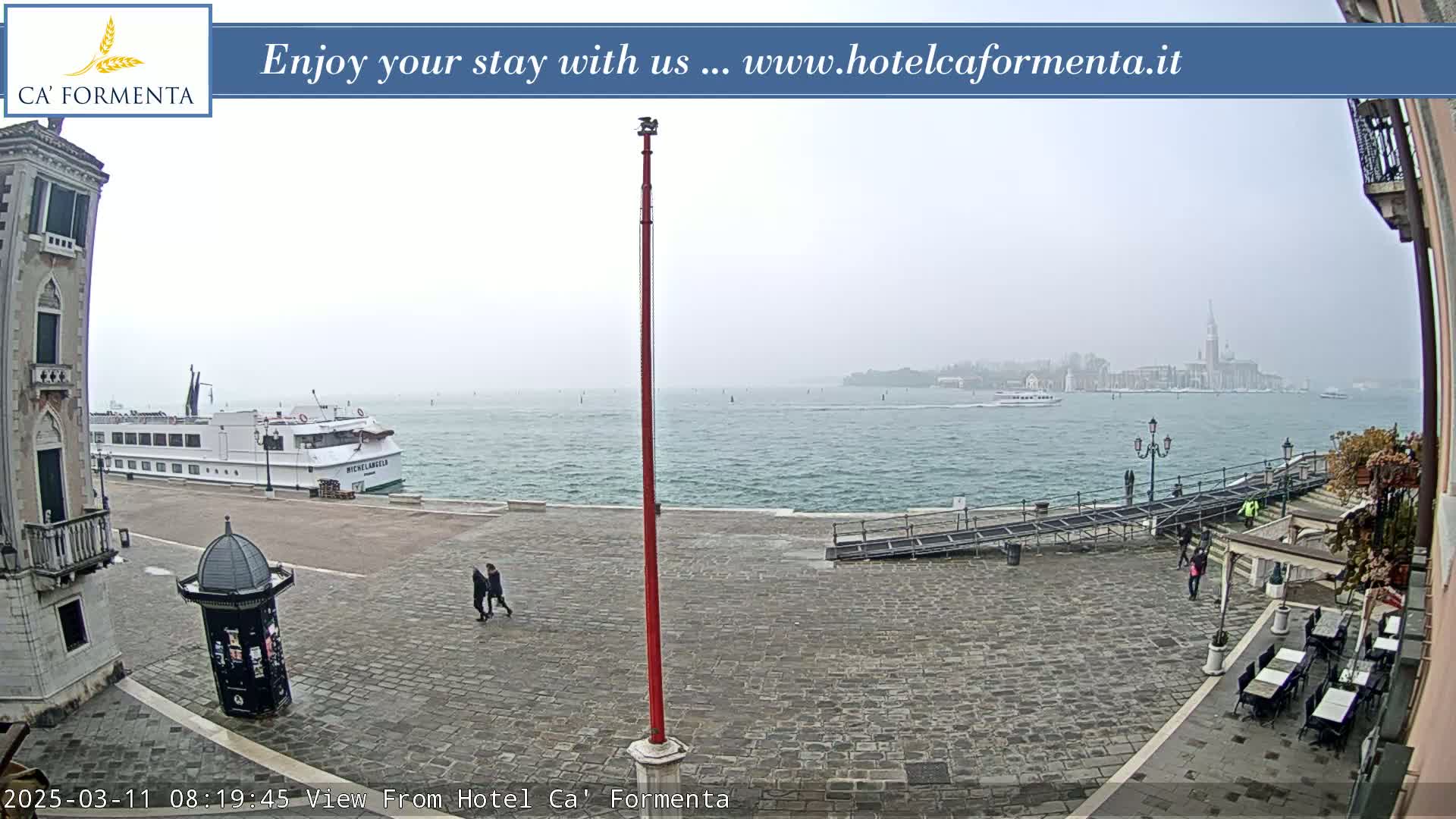 A hazy view of a waterway shows a large passenger ship docked near a paved waterfront area with a few pedestrians and outdoor seating; in the distance, a city skyline is visible under a foggy sky.