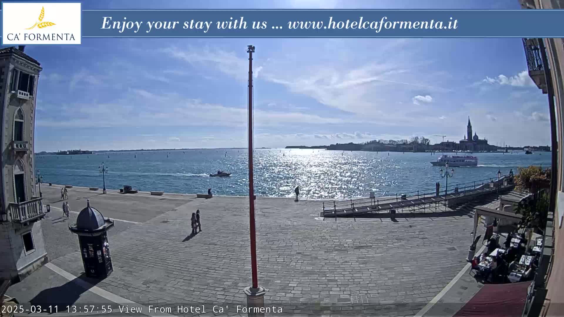 A sunny day overlooking a calm body of water with a boat and distant buildings, and a paved plaza with a few people.