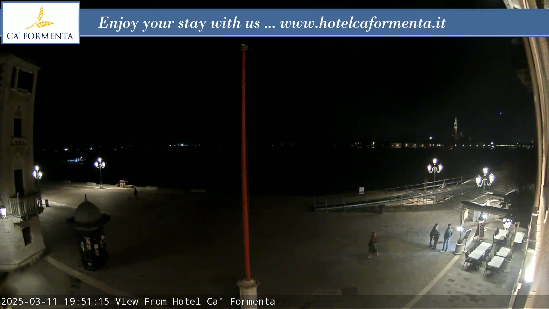 A nighttime view of a plaza overlooking a dark body of water with lights across the far shore, under a clear sky.