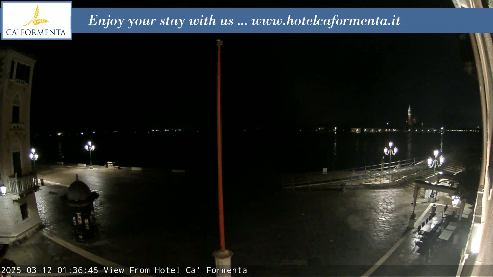 A nighttime view of a cobblestone plaza and waterfront, illuminated by lampposts, with calm dark water and distant lights across the water.