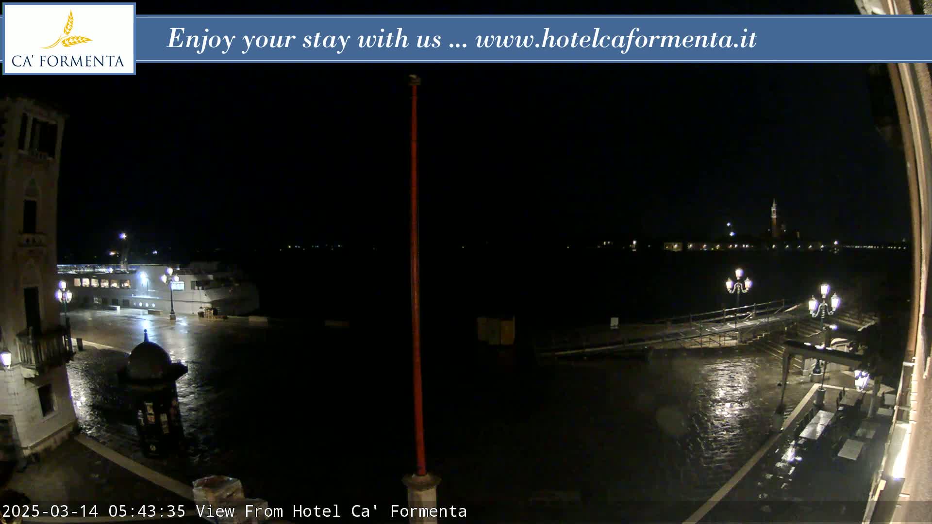 A nighttime view of a waterfront area showing a wet, paved plaza, a docked boat, and distant city lights across the water.