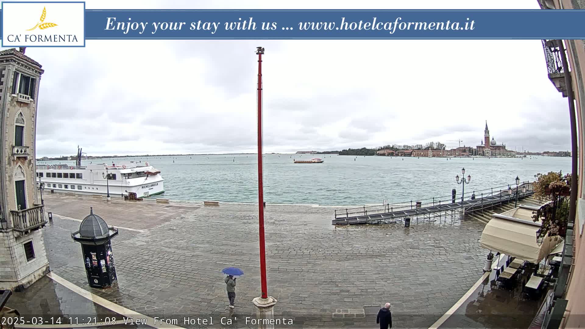 A wet, paved waterfront area overlooks a calm body of water with a cruise ship and distant buildings under an overcast sky.