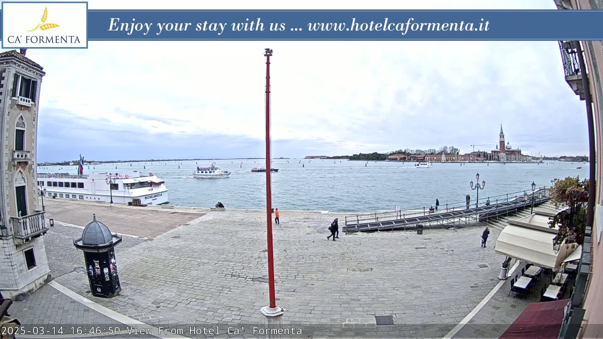 A paved plaza overlooks a waterway with boats and a distant cityscape under an overcast sky.