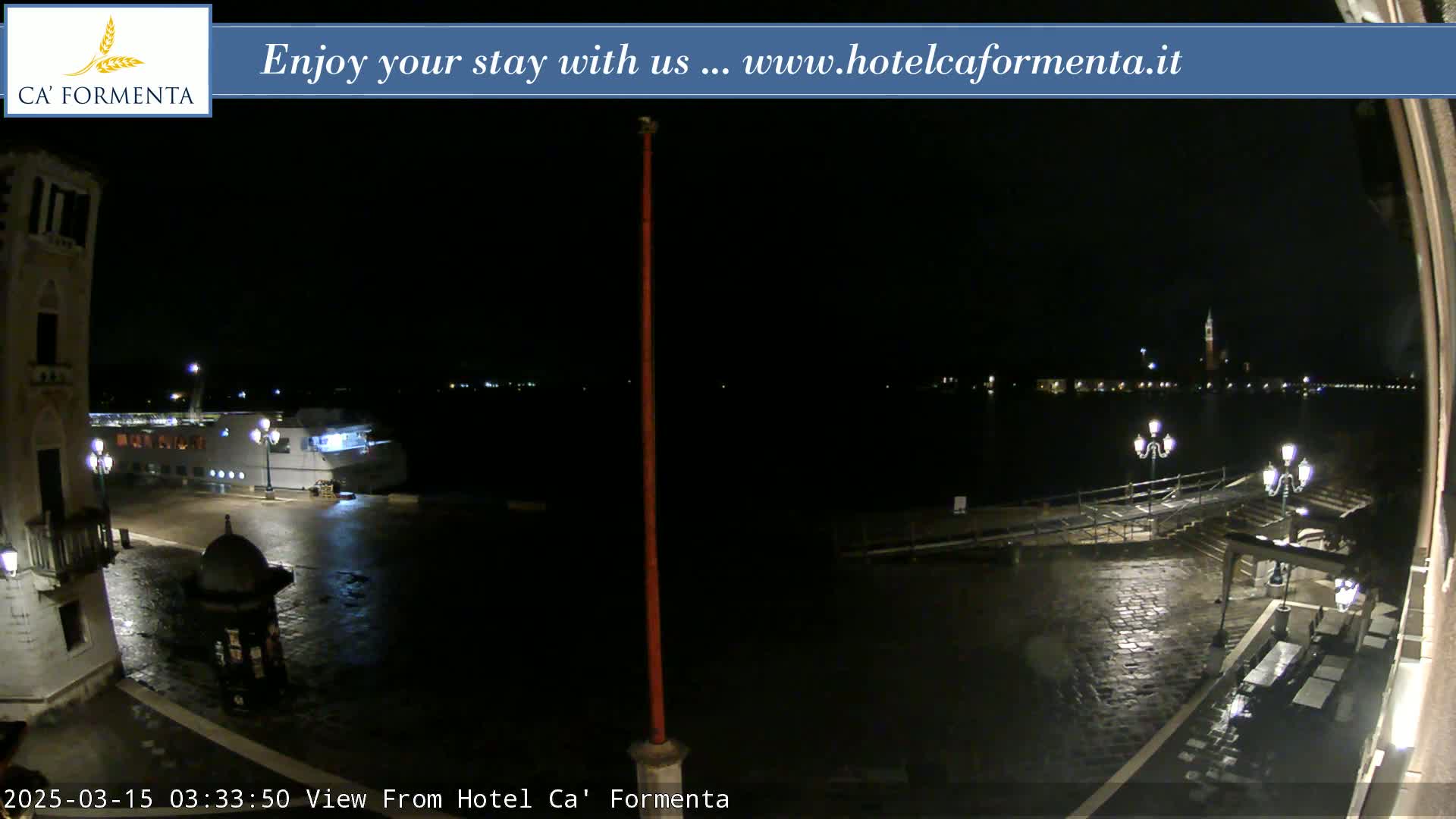 A nighttime view of a wet waterfront plaza with a docked boat, some distant lights, and a building to the left.