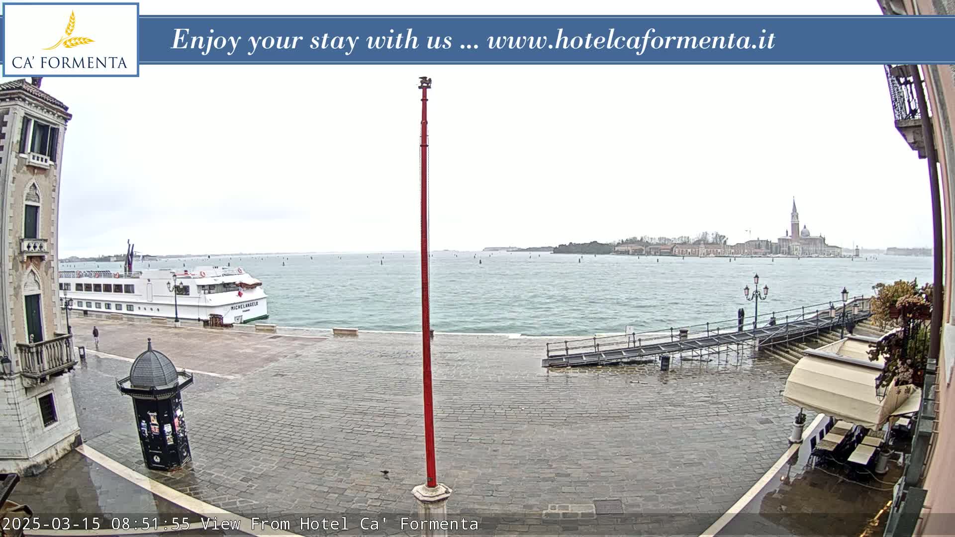 A wet, paved waterfront area shows a large passenger ship and a distant island cityscape under an overcast sky.