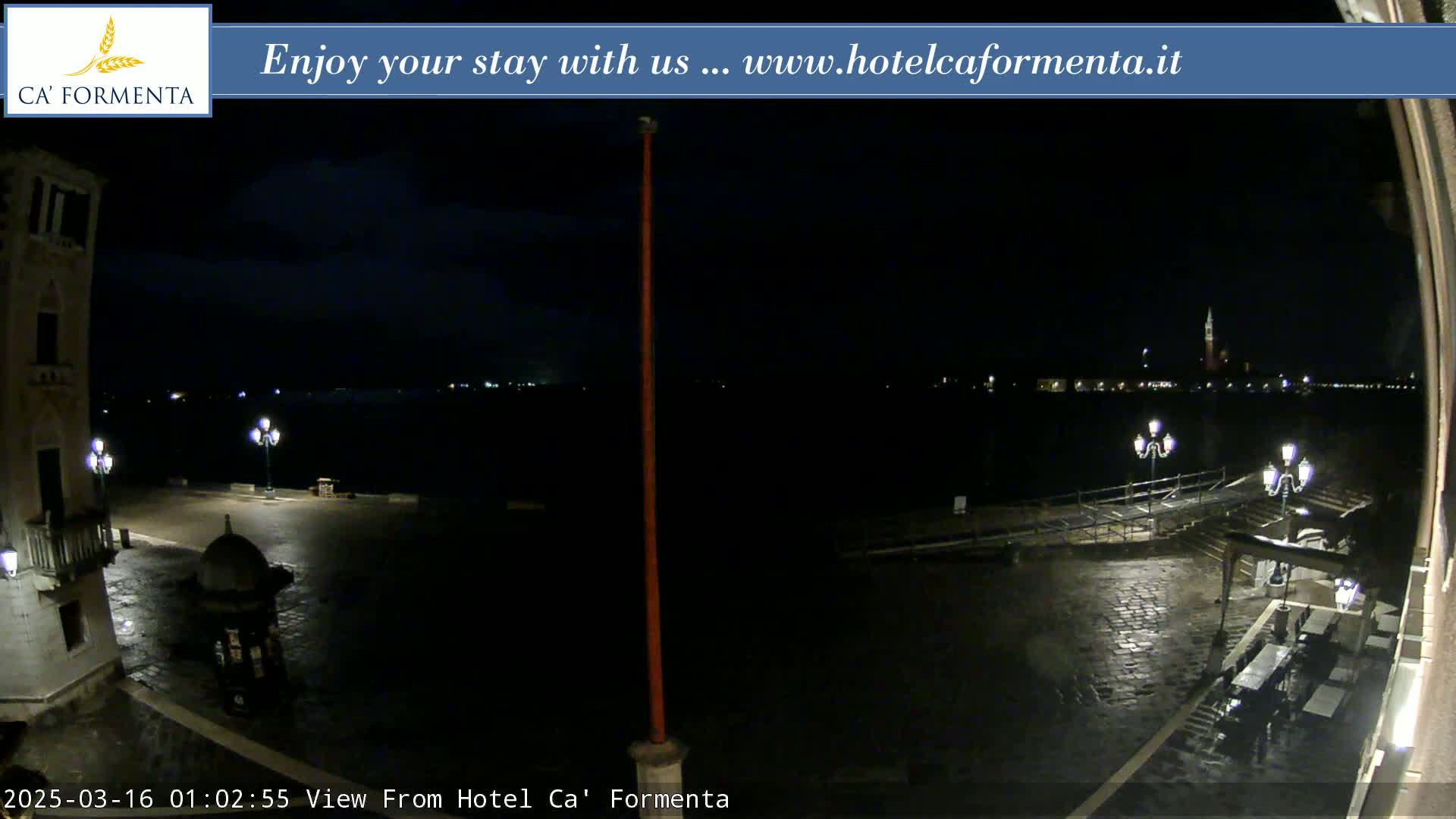 A dark, nighttime view of a cobblestone plaza overlooking a calm body of water, with distant city lights visible across the water.