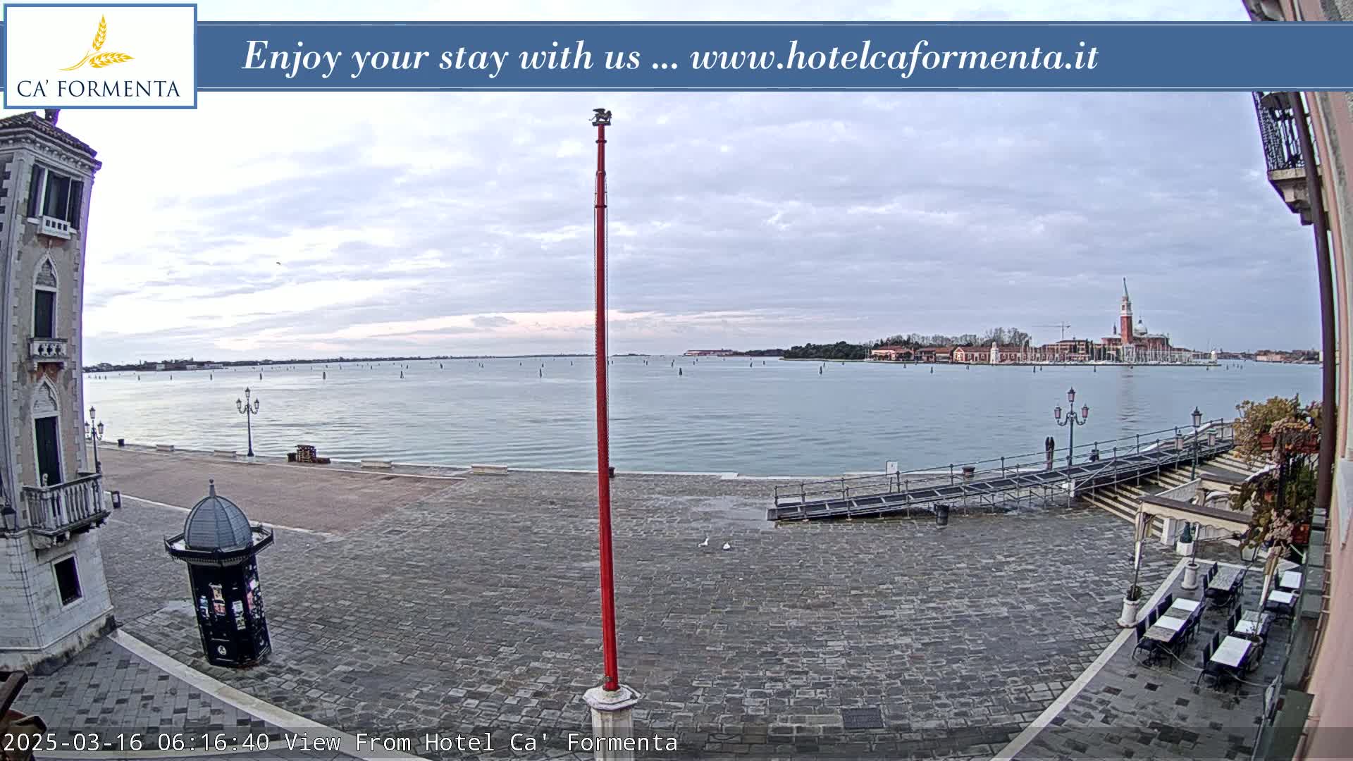 A cobblestone plaza overlooks calm water, with a distant island and overcast skies.