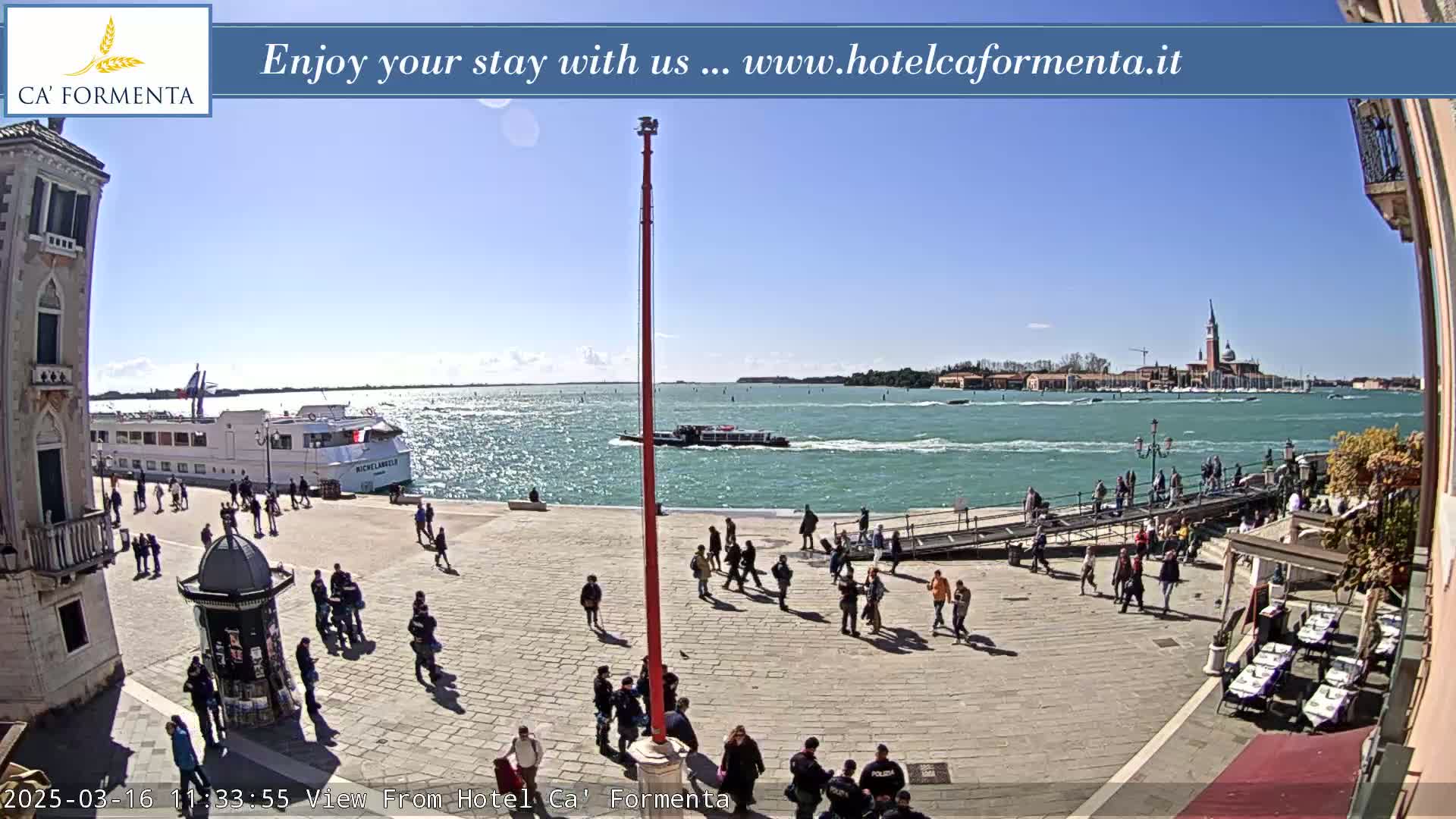 A sunny day shows a waterfront plaza with people walking, boats on the water, and a distant island with buildings.