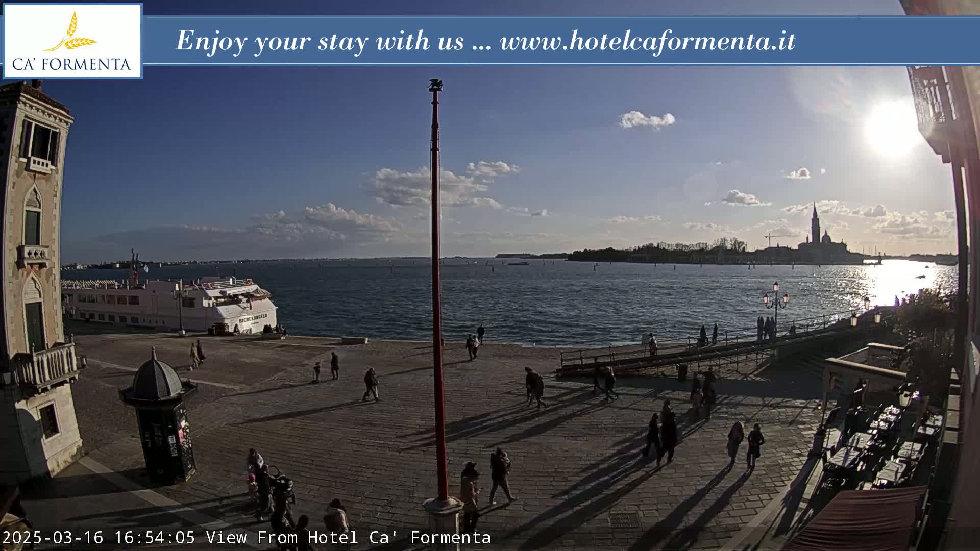 A sunny waterfront plaza with scattered pedestrians, a large cruise ship, and a distant church visible across the water.