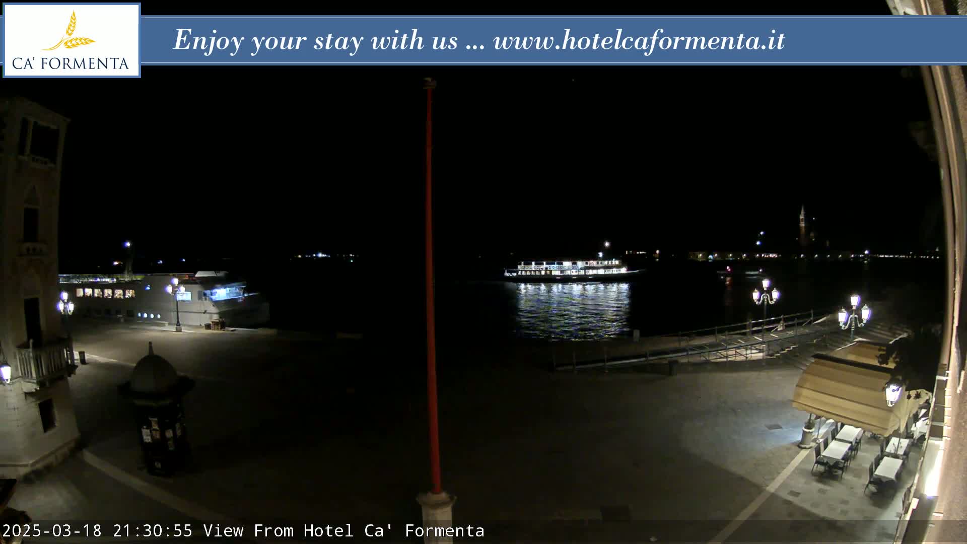 A nighttime view of a waterfront plaza with a boat on the water, illuminated buildings in the distance, and outdoor seating at a restaurant under a retractable awning.