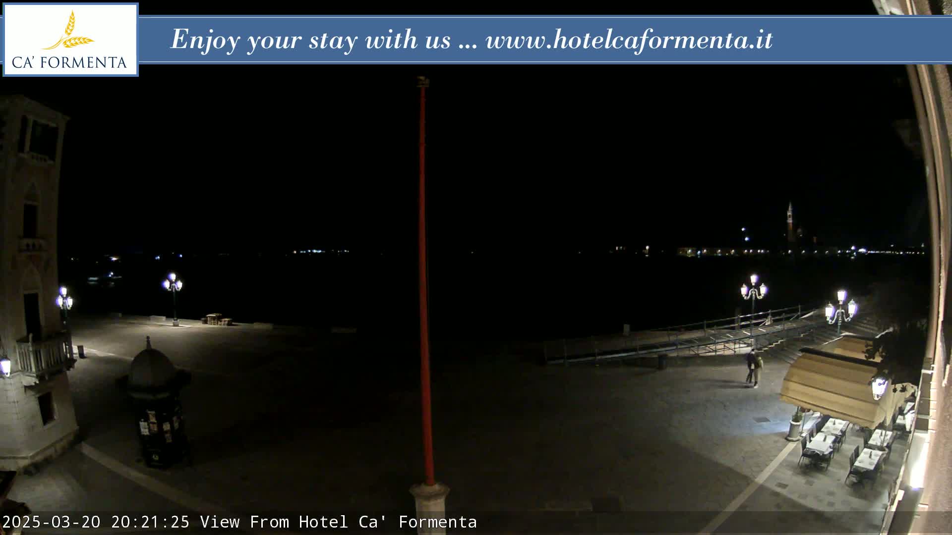 A nighttime view of a waterfront plaza with a few people and outdoor seating at a restaurant, under a dark sky.