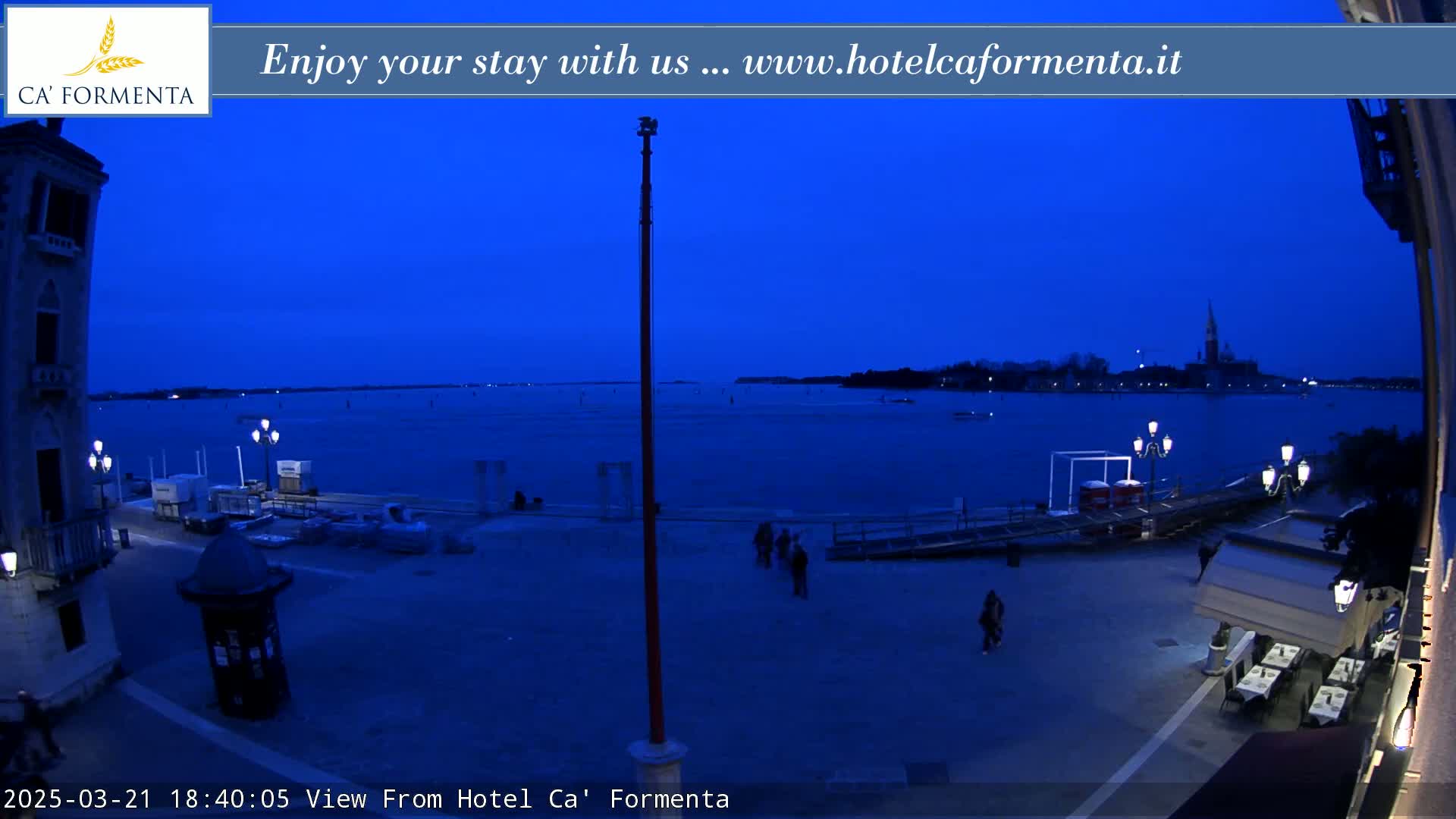 A twilight view shows a waterfront plaza with a few people, some boats, and buildings across a calm body of water.