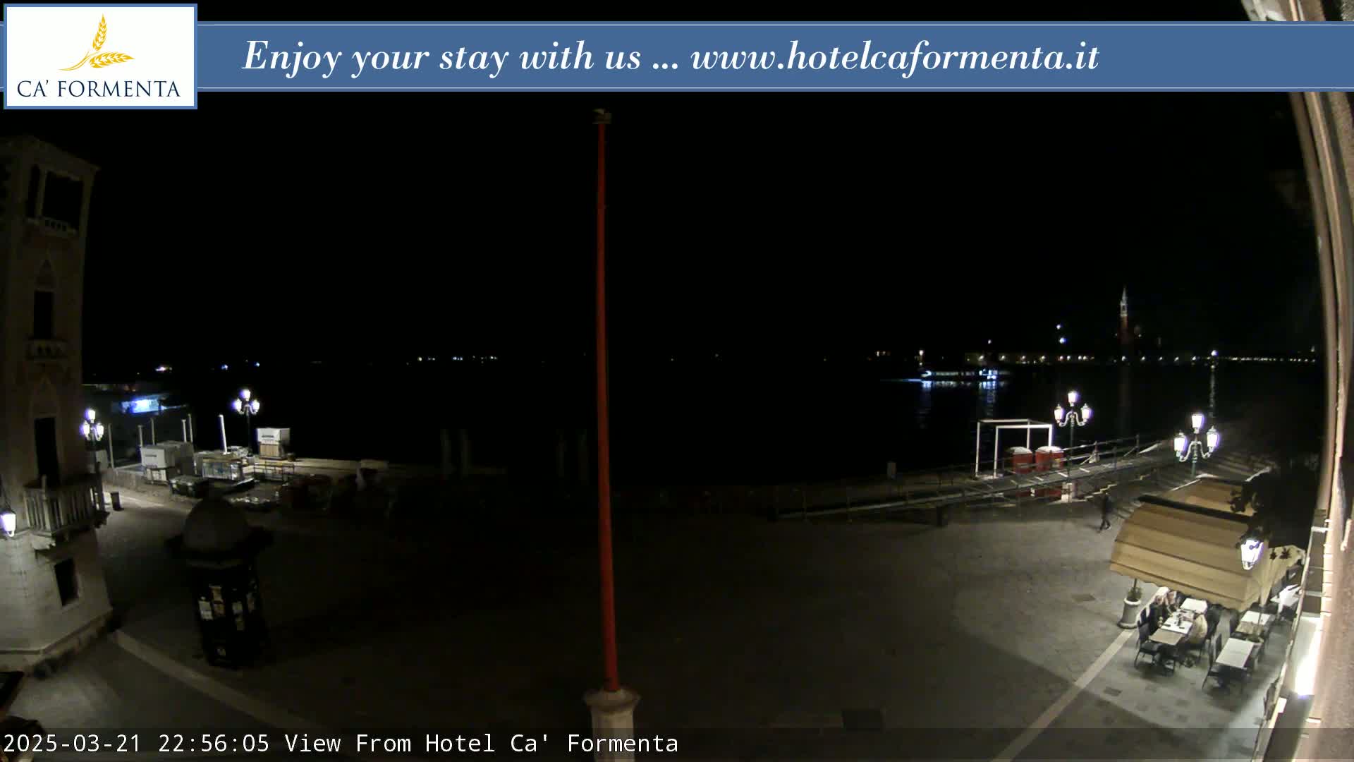 A nighttime view of a waterfront plaza with a calm body of water reflecting lights across from a building, and a few people dining outdoors at a restaurant.