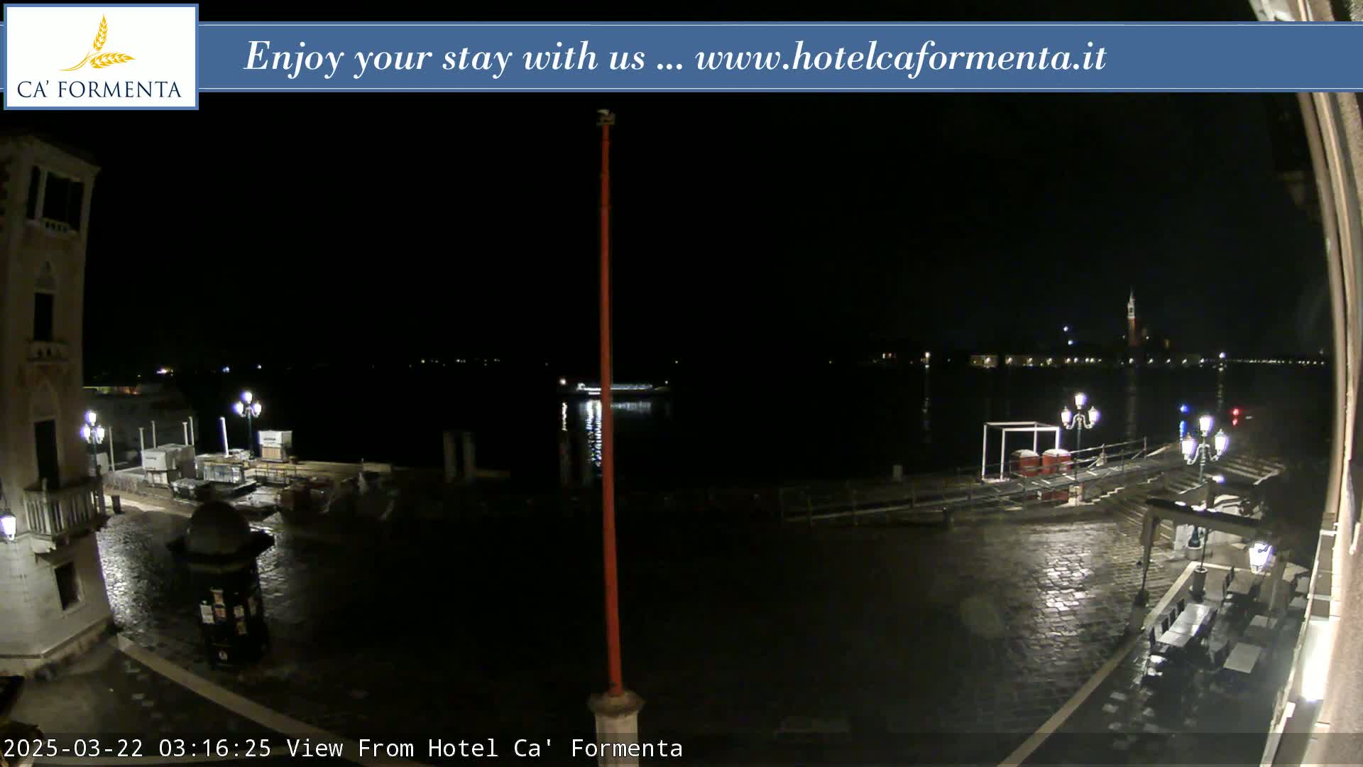 A nighttime view of a waterfront plaza, wet from rain, shows a dark body of water with lights across it, and buildings along the shore.
