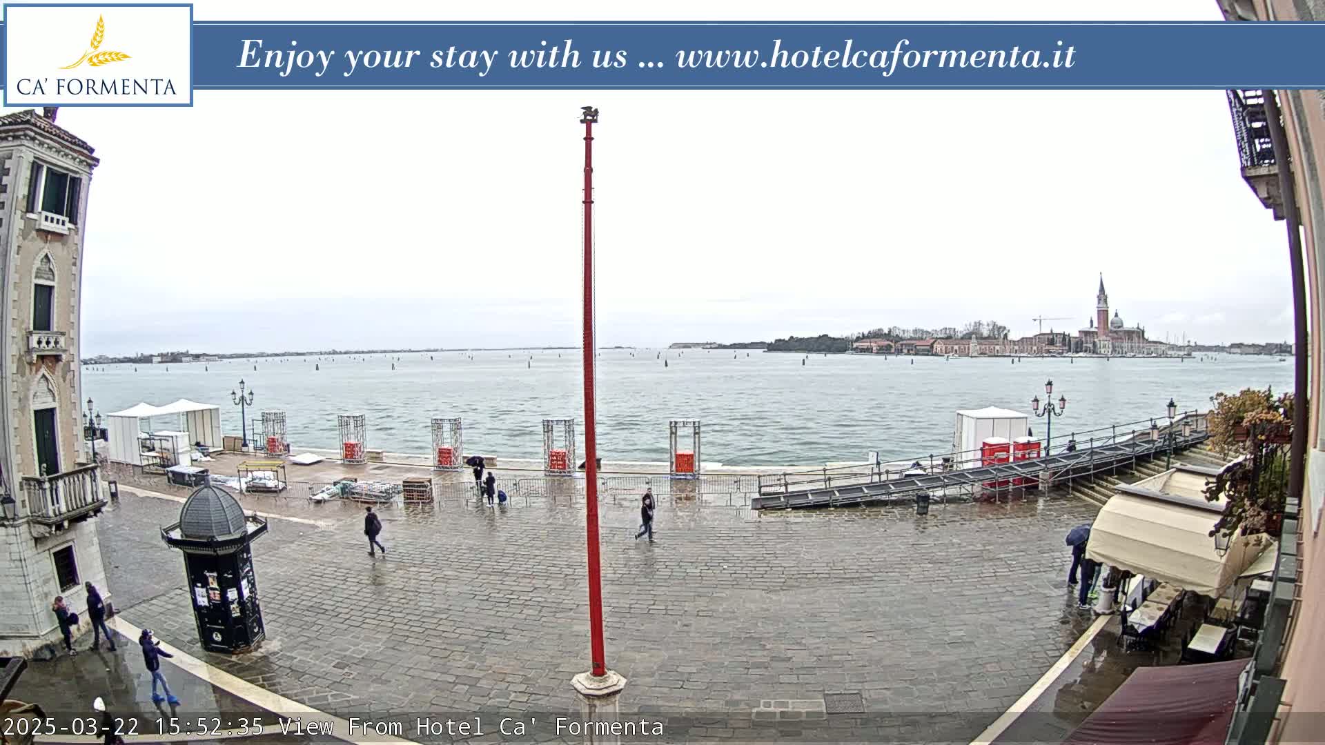 A wet cobblestone plaza overlooks a calm body of water with a distant city skyline under an overcast sky.