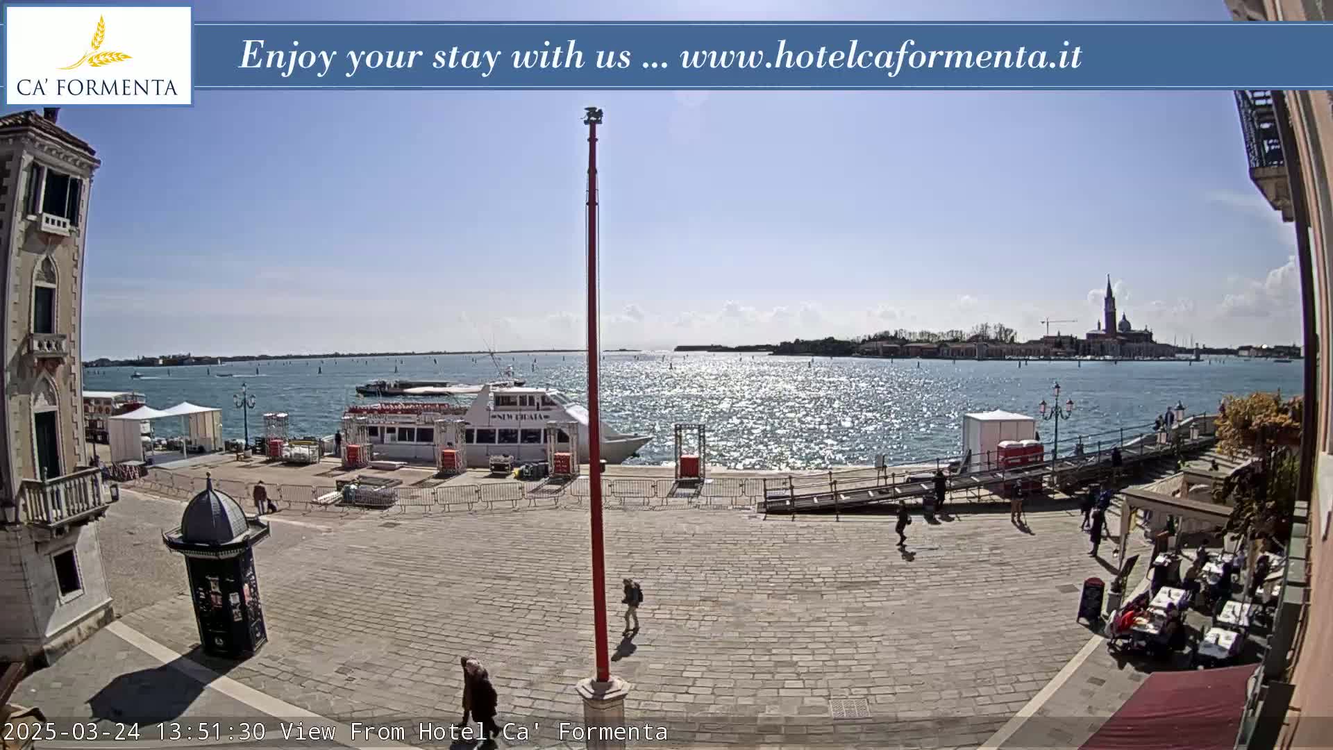 A sunny waterfront plaza scene shows a docked boat, several people walking, and a distant island with buildings under a clear sky.