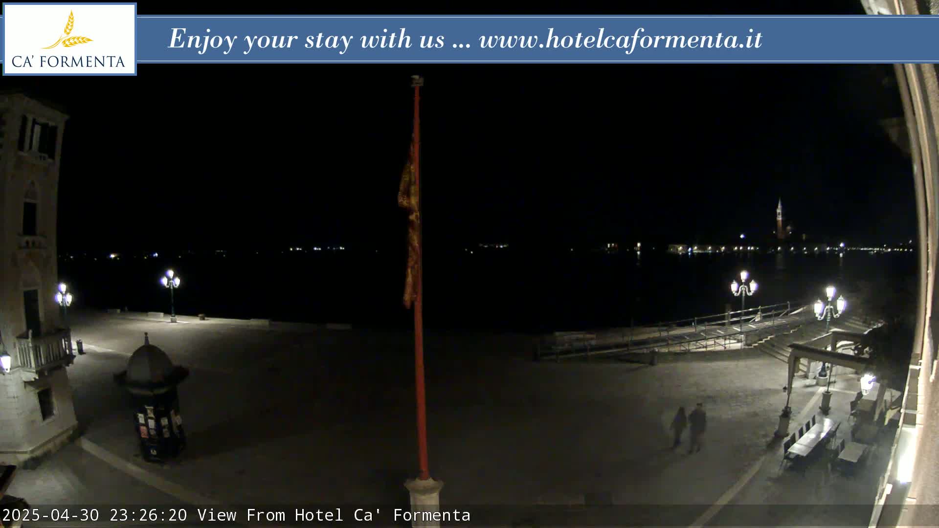 A nighttime view of a plaza overlooking a calm body of water with lights illuminating the far shore and a few people walking in the foreground.