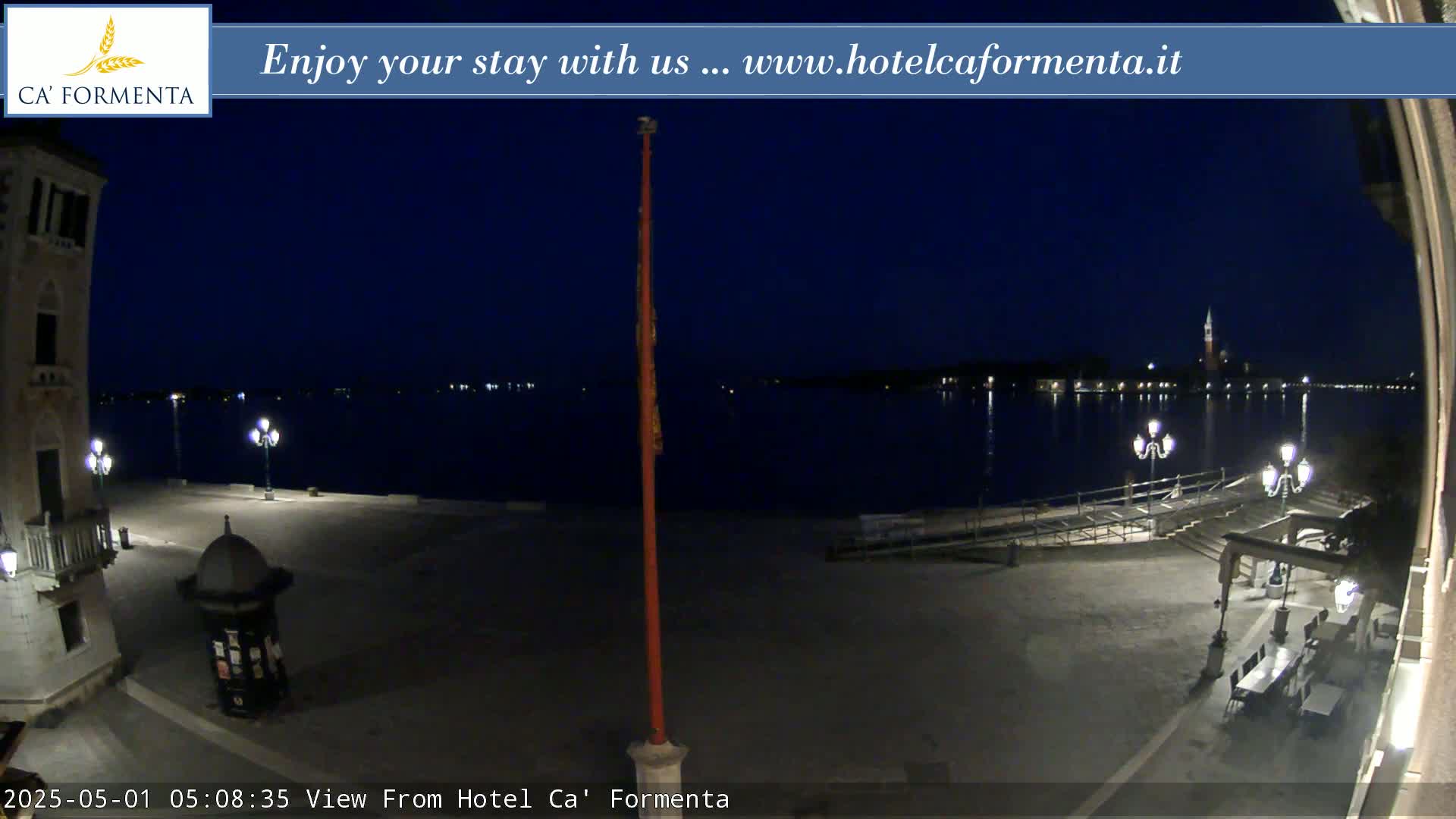 A nighttime view of a calm body of water, illuminated by lights along the shore and across the water, with a paved plaza and building visible in the foreground.