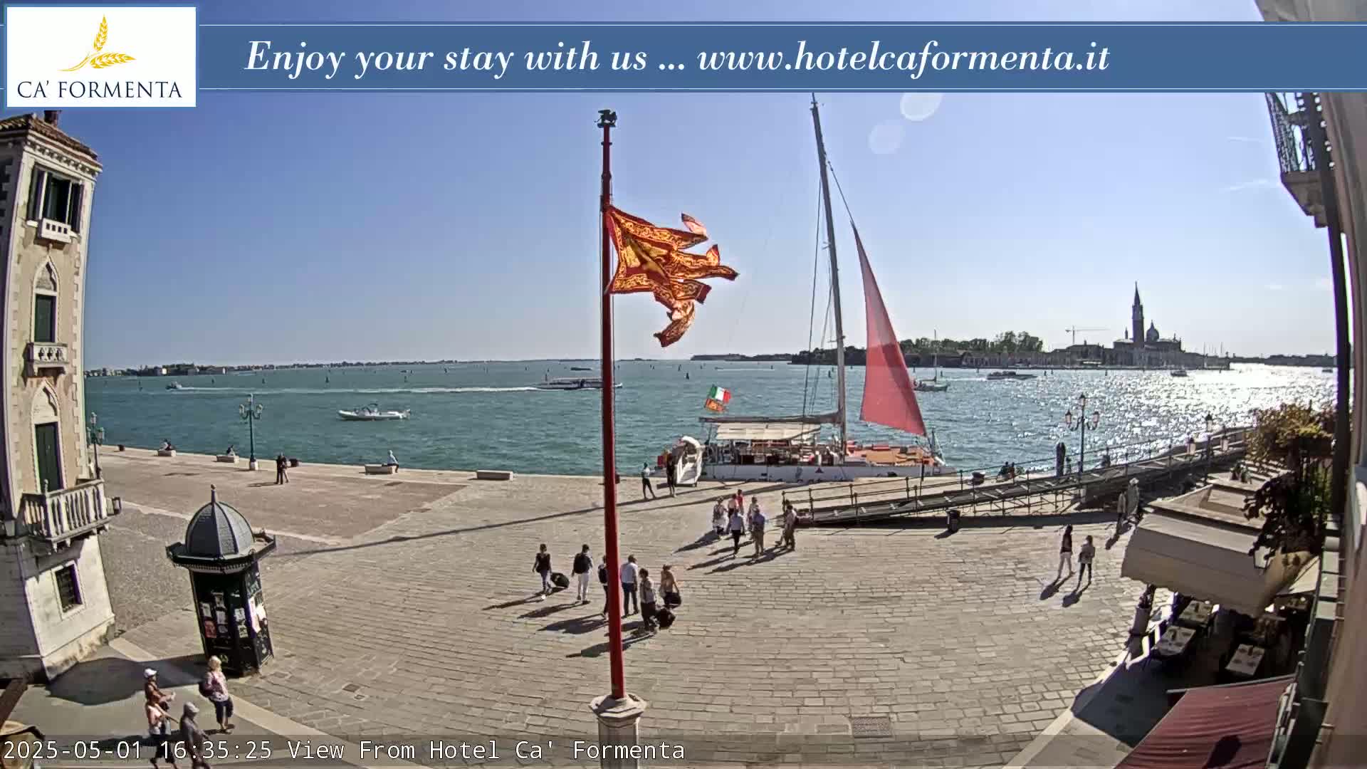 A sunny day shows a paved plaza overlooking a calm body of water with boats and a sailboat, and several people are walking around the plaza.