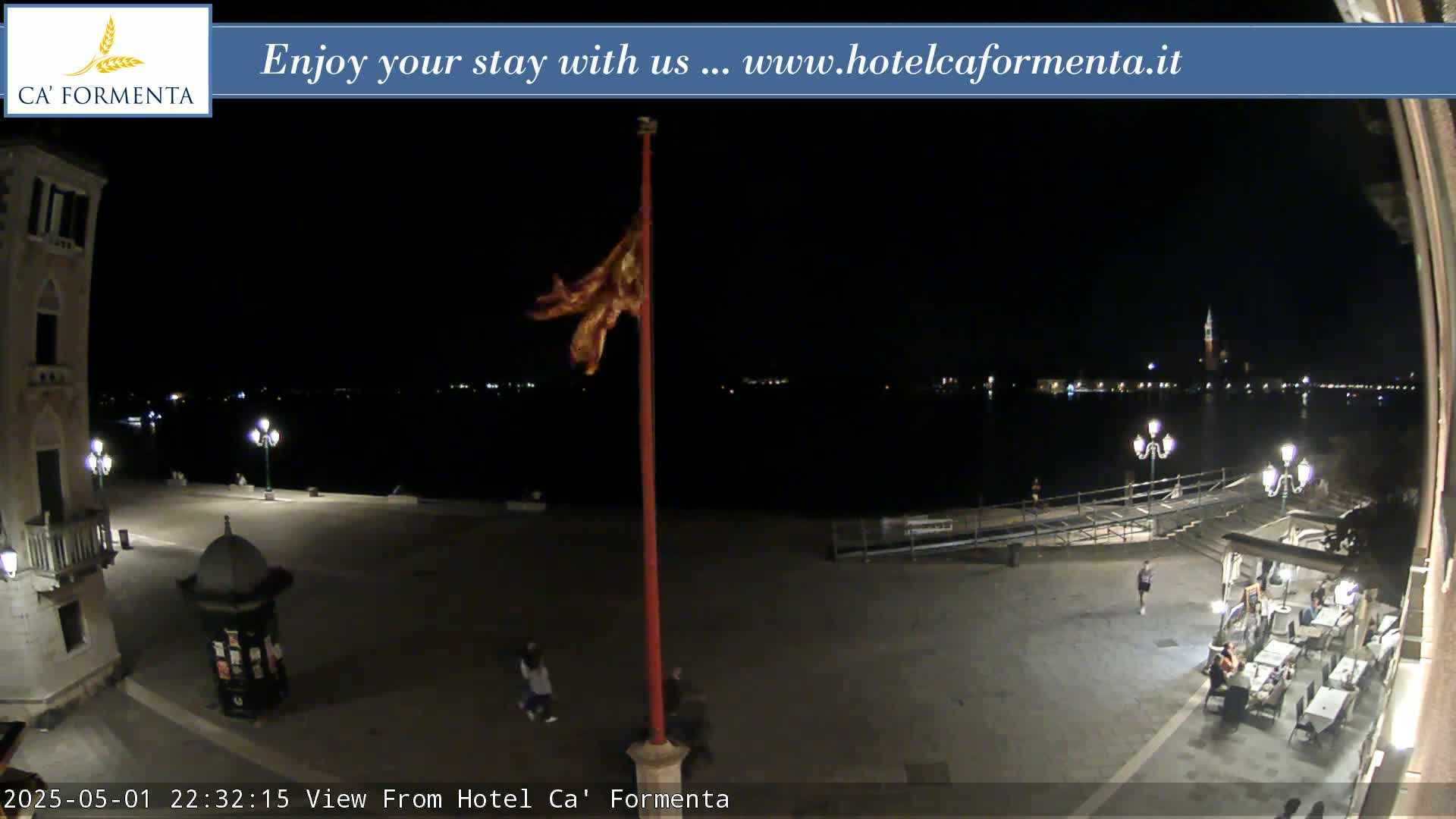 A nighttime view of a plaza overlooking a calm body of water with city lights in the distance, a flagpole with a flag waving slightly, and a few people in the plaza.