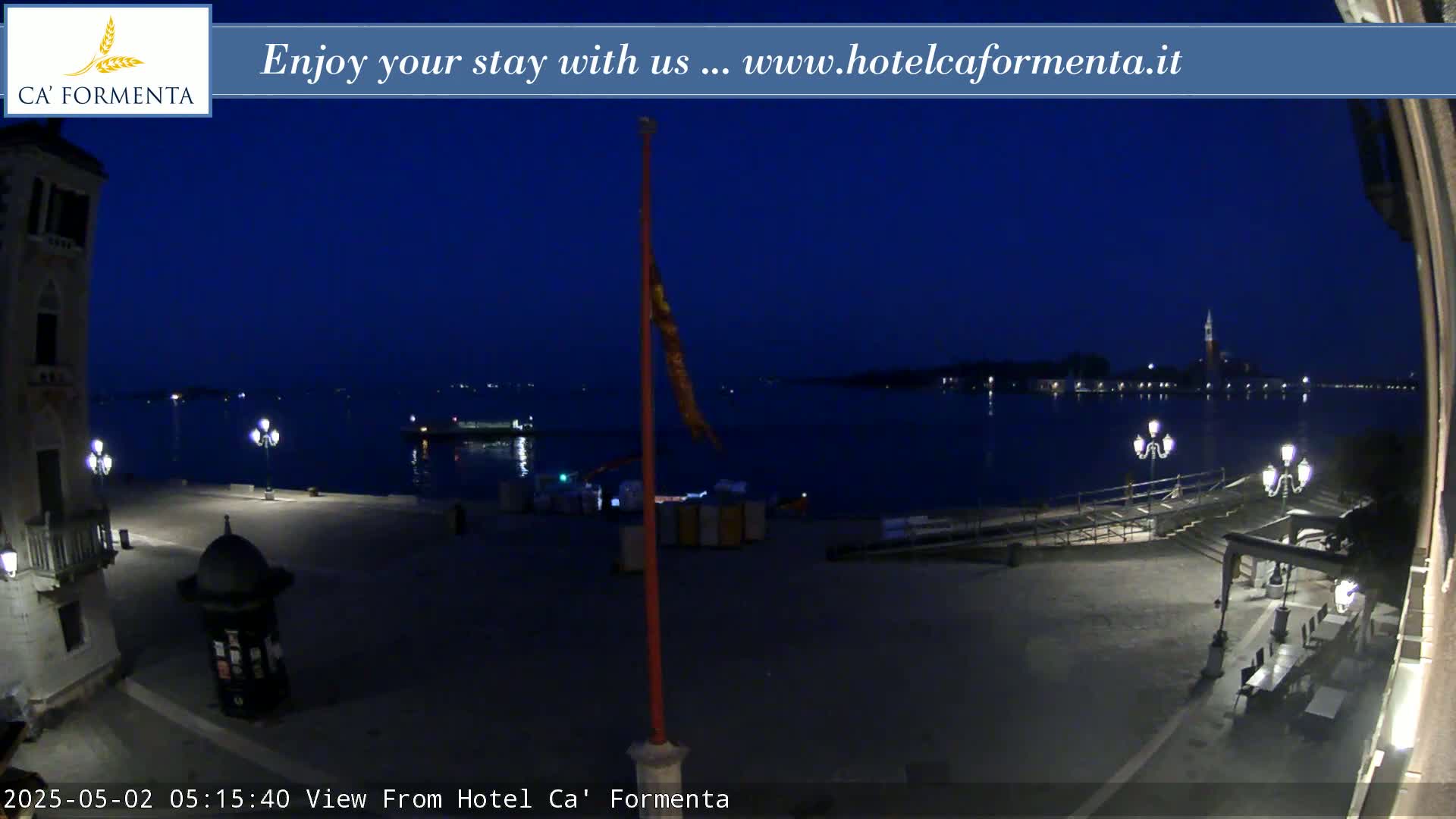 A nighttime view of a waterfront plaza, showing a calm body of water with distant lights across it under a dark blue sky.