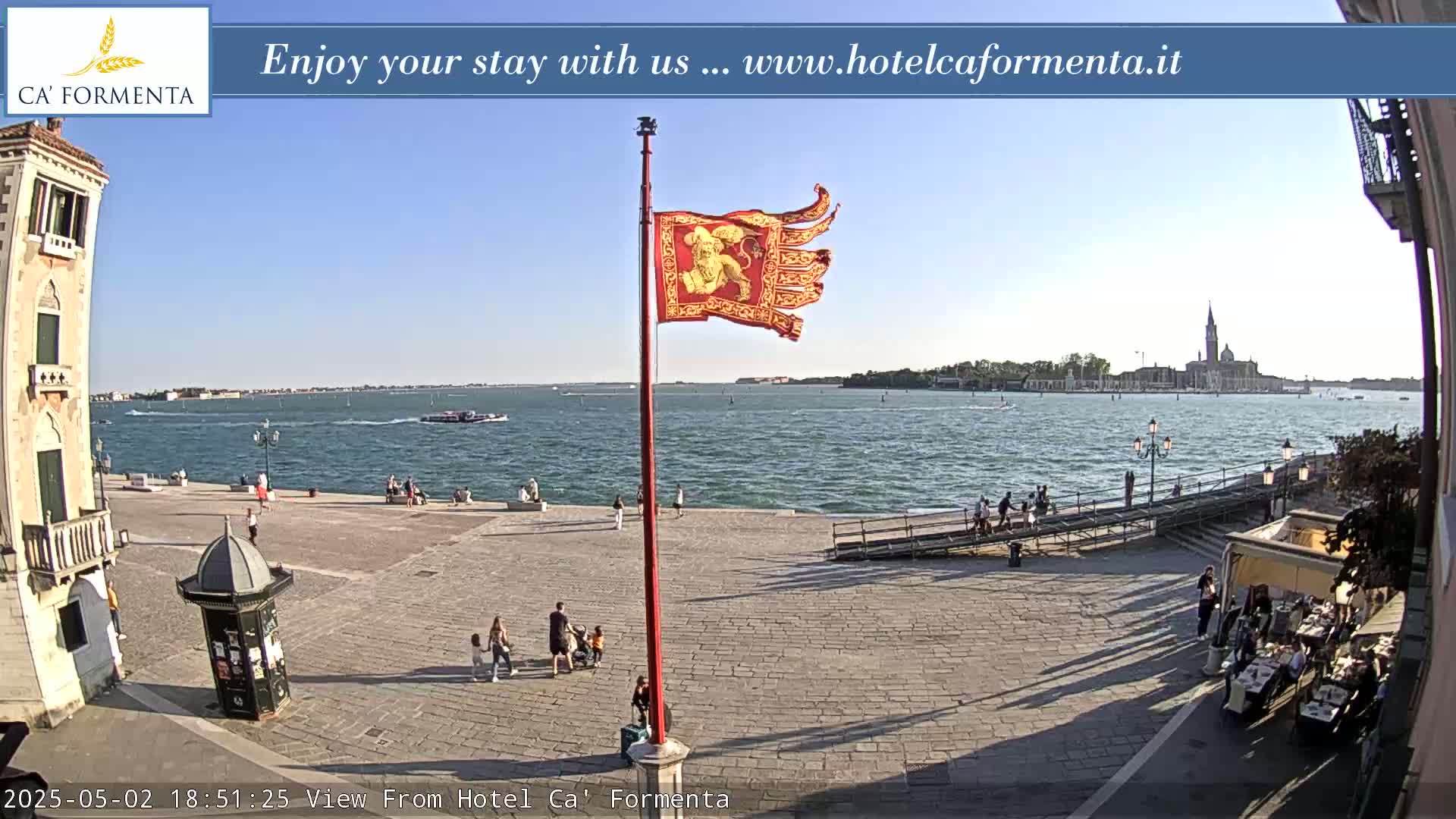 A sunny Venetian waterfront square shows a red flag with a lion on it, people strolling, and a body of water with distant buildings and boats.