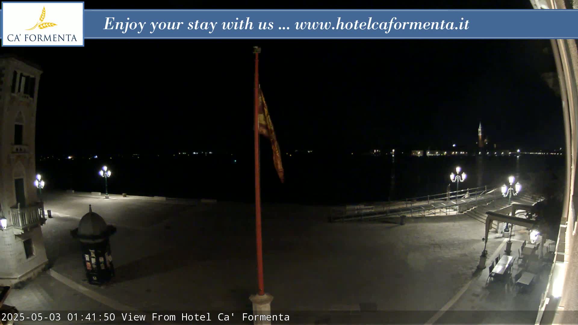 A nighttime view of a plaza overlooking calm water, with a flagpole and distant lights visible across the water.