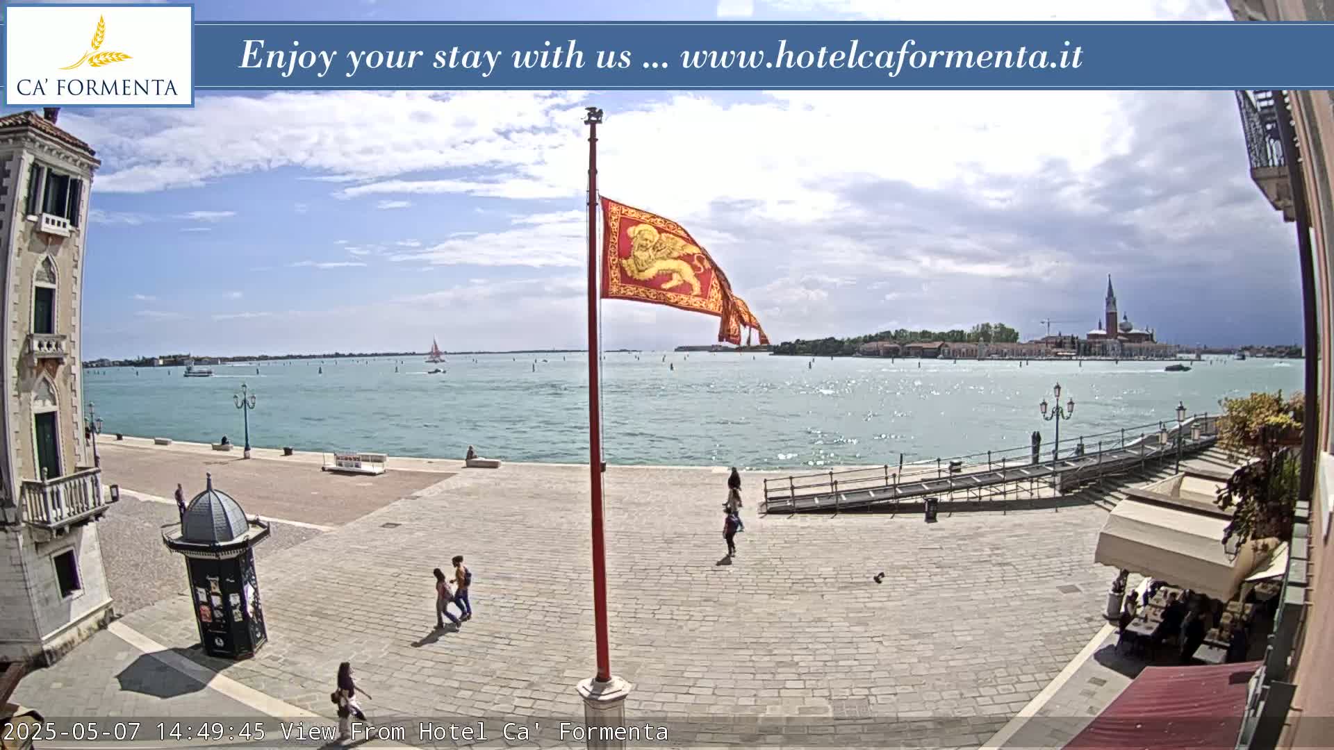 A partly cloudy day shows a Venetian waterfront square with a red flag bearing a lion, people walking, and a view of the water and distant buildings.