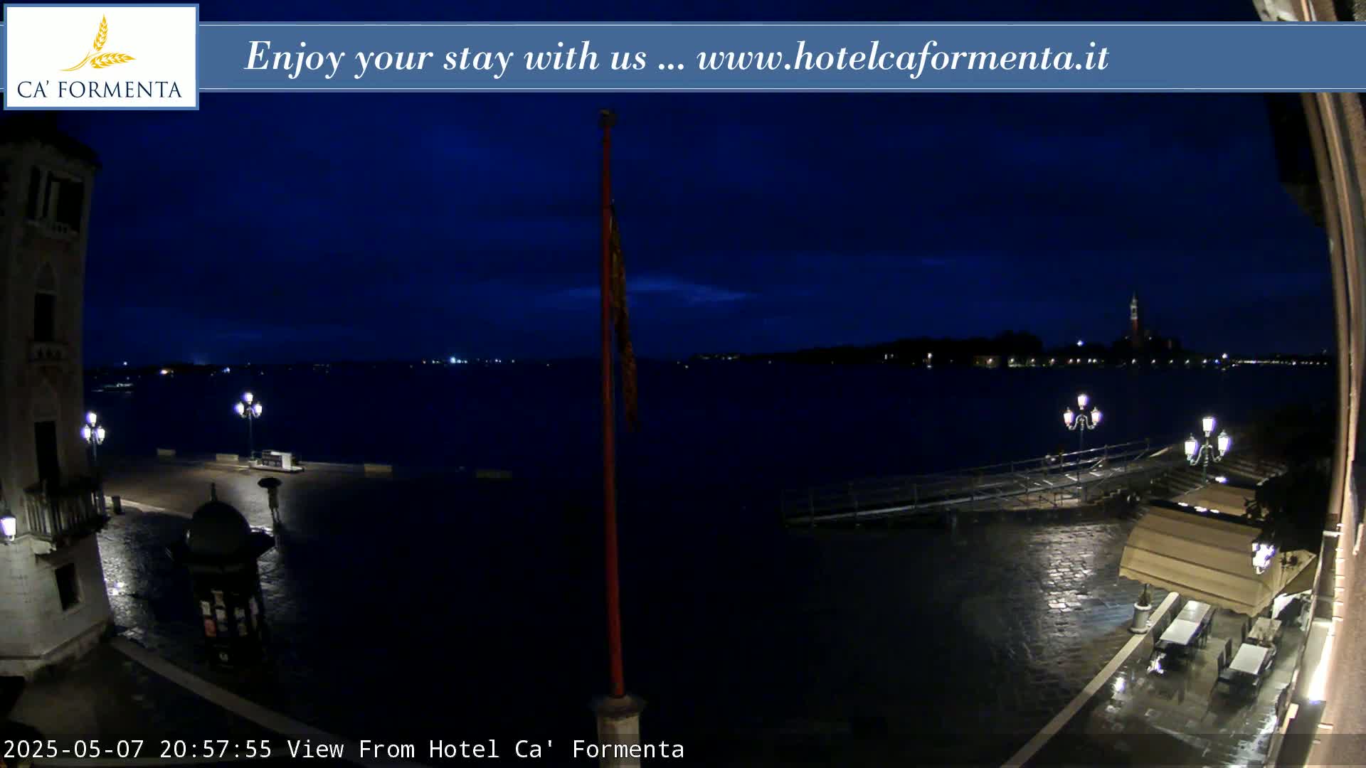 A nighttime view of a waterfront plaza shows a dark body of water under a twilight sky, with wet paving stones reflecting lights from street lamps and a restaurant with outdoor seating.