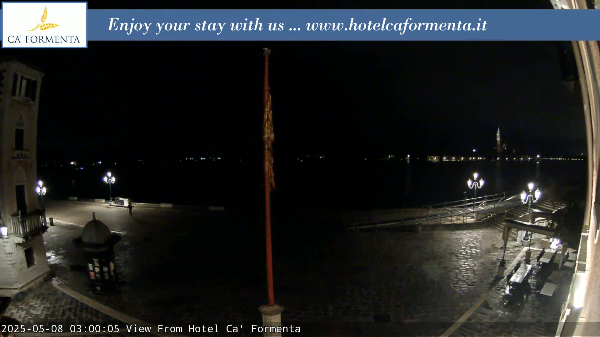 A nighttime view of a cobblestone plaza and waterfront, showing dimly lit buildings and a calm body of water with lights visible across it.