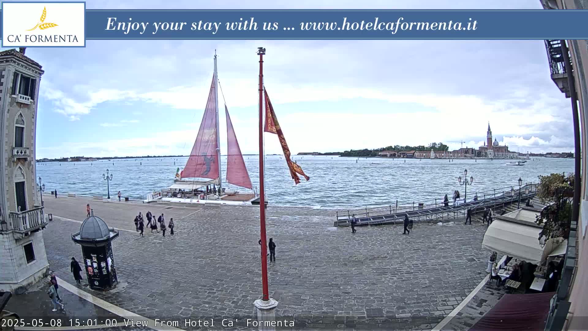 A waterfront view shows a sailboat docked near a stone plaza with people walking, under a mostly cloudy sky, with a distant island visible across the water.