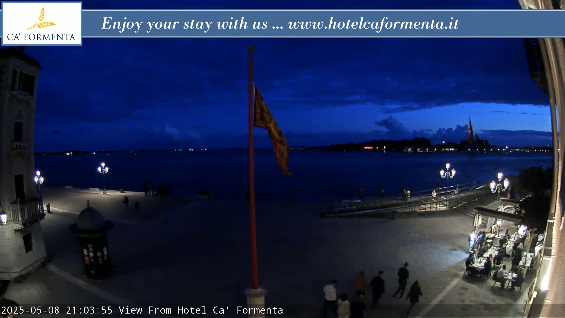 A nighttime view of a waterfront plaza, showing a calm body of water with city lights in the distance under a dark blue, mostly clear sky.