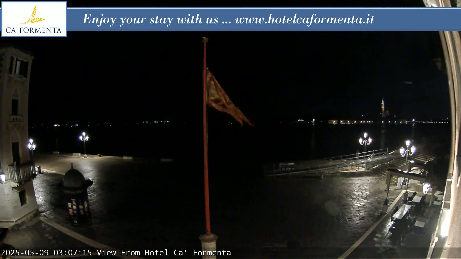 A nighttime view of a plaza overlooking a dark body of water, with a flagpole and faint city lights in the distance.