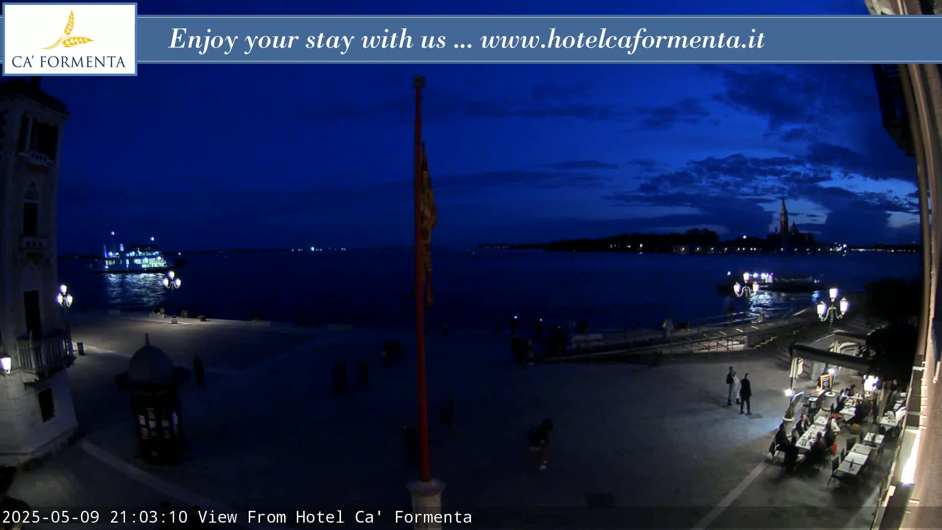 A nighttime view of a waterfront plaza with a few people, boats on the water, and distant city lights under a dark blue sky.