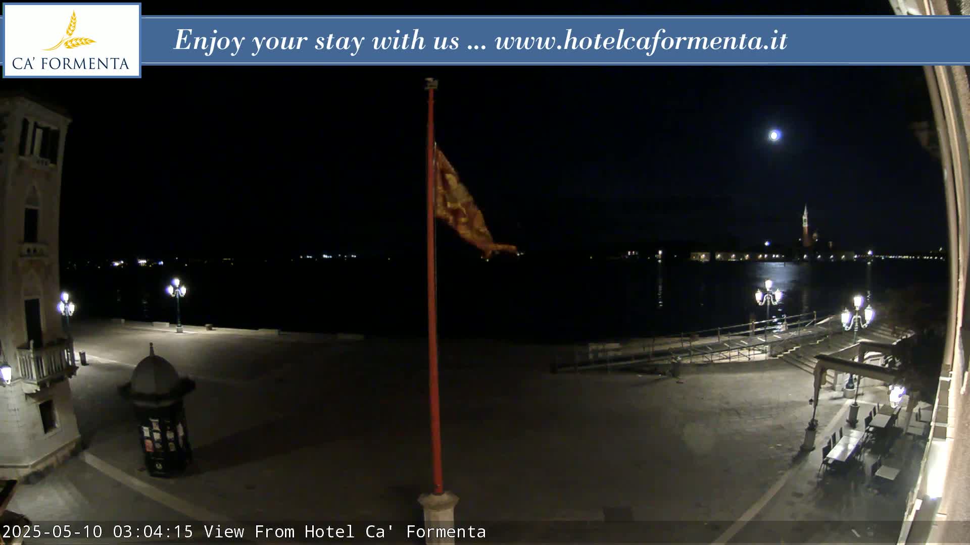 A nighttime view of a calm body of water, under a full moon, shows a flagpole with a flag and some outdoor furniture in a plaza near a building.