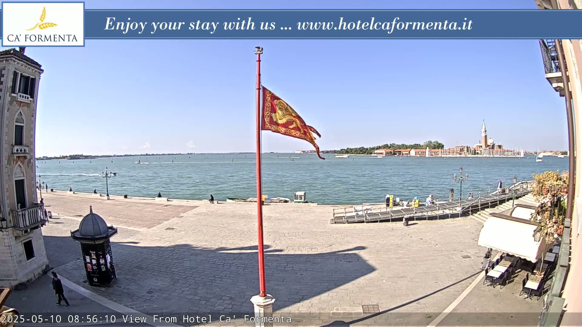 A sunny waterfront view shows a stone plaza with a flagpole and a distant island.
