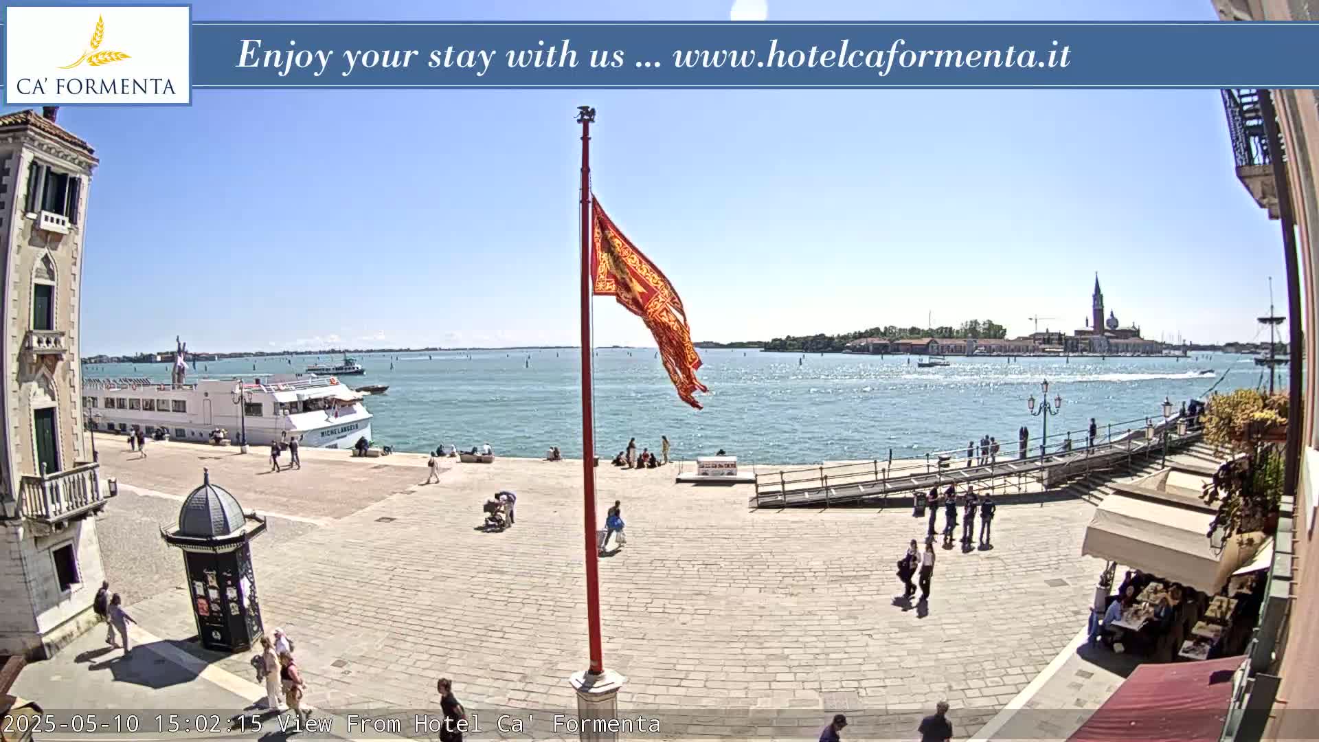 A sunny waterfront plaza shows a large cruise ship, a red flag on a pole, and several people, with a distant view of buildings and water.