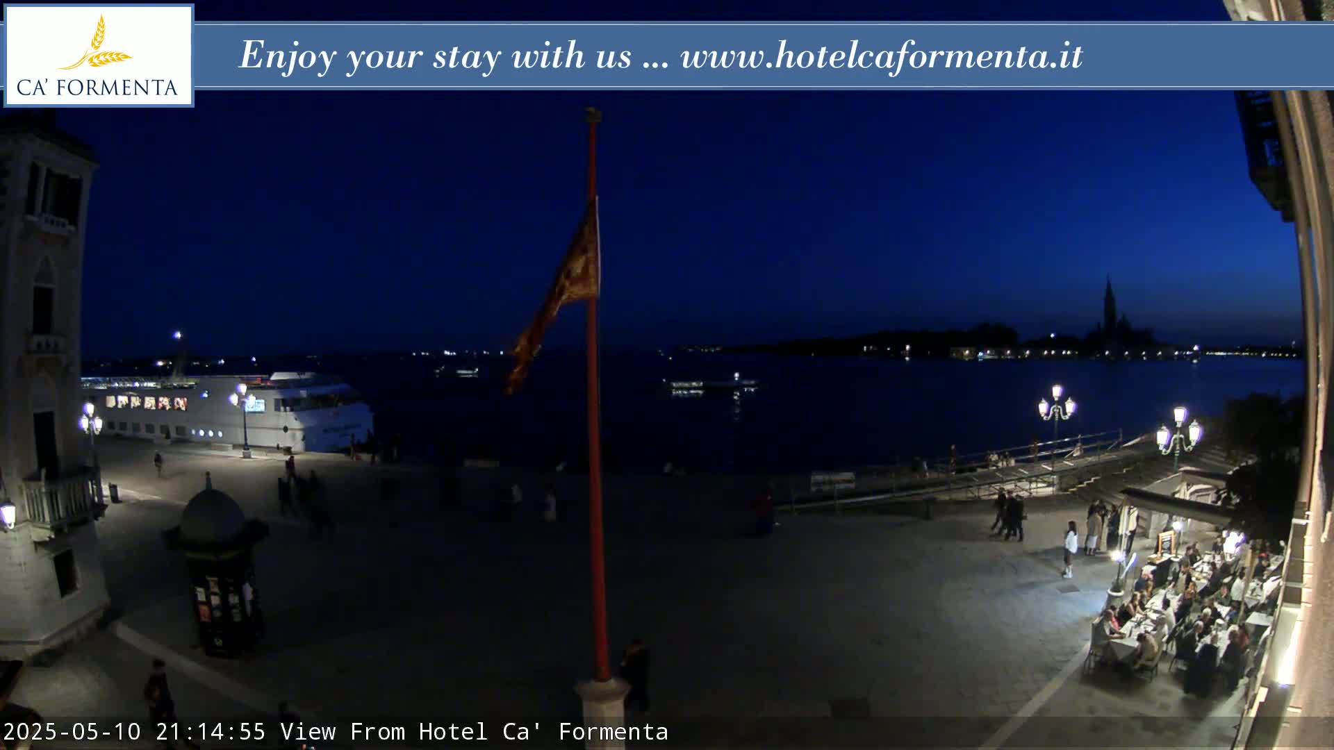 A nighttime view of a waterfront plaza with a restaurant, a docked boat, and a flagpole, under a dark blue twilight sky.