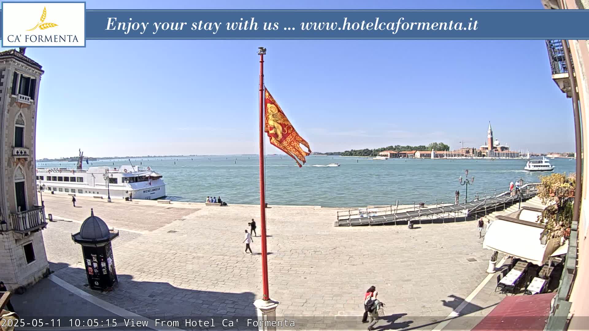 A sunny waterfront plaza scene shows a large passenger ship docked near a flagpole with a red flag featuring a lion, several people walking around, and a distant view of a church on an island.