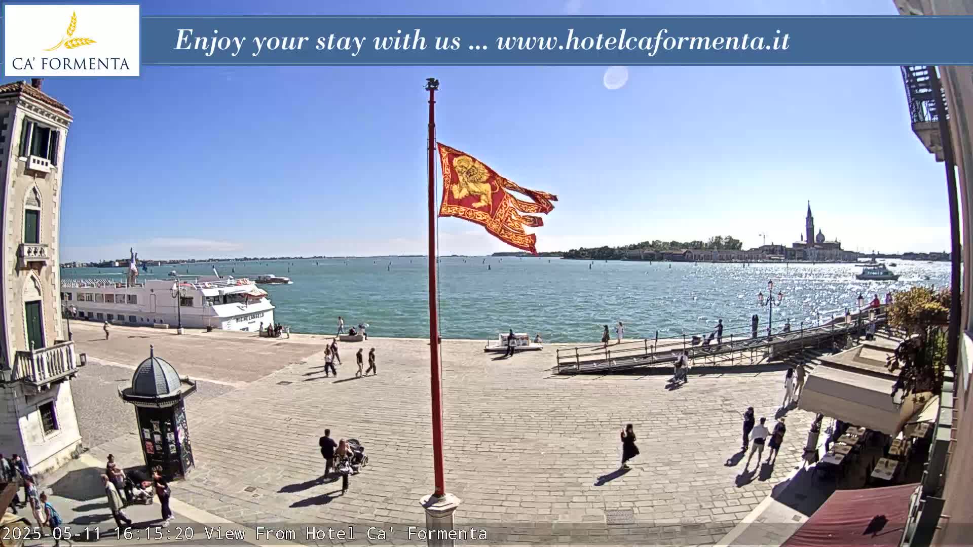 A sunny waterfront plaza scene shows a large vessel in the distance, several people walking, and a tattered red flag with a lion emblem waving on a tall pole.