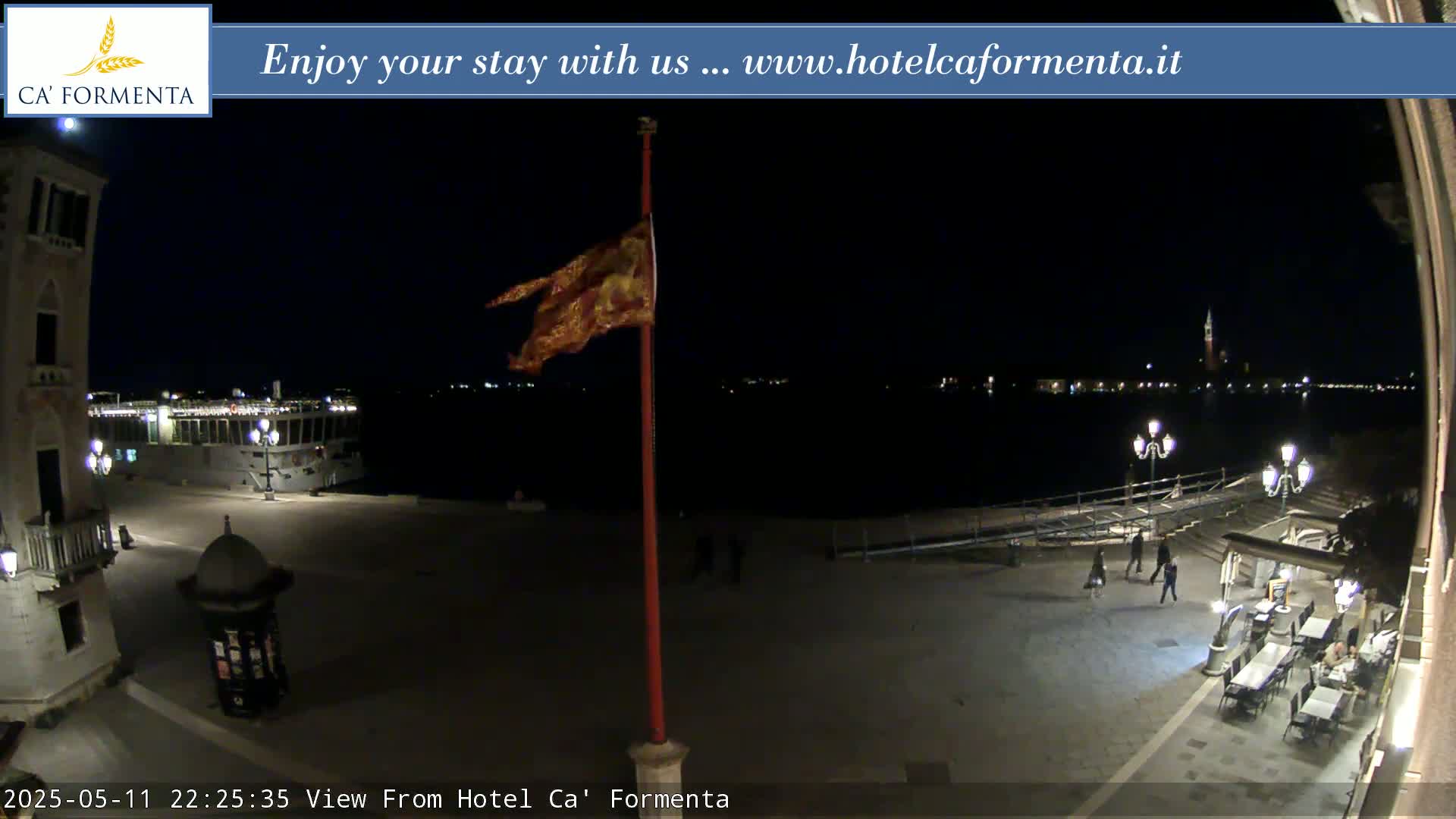 A nighttime view of a waterfront plaza shows a flag waving in the breeze, a few people walking, and a body of water with lights across it.