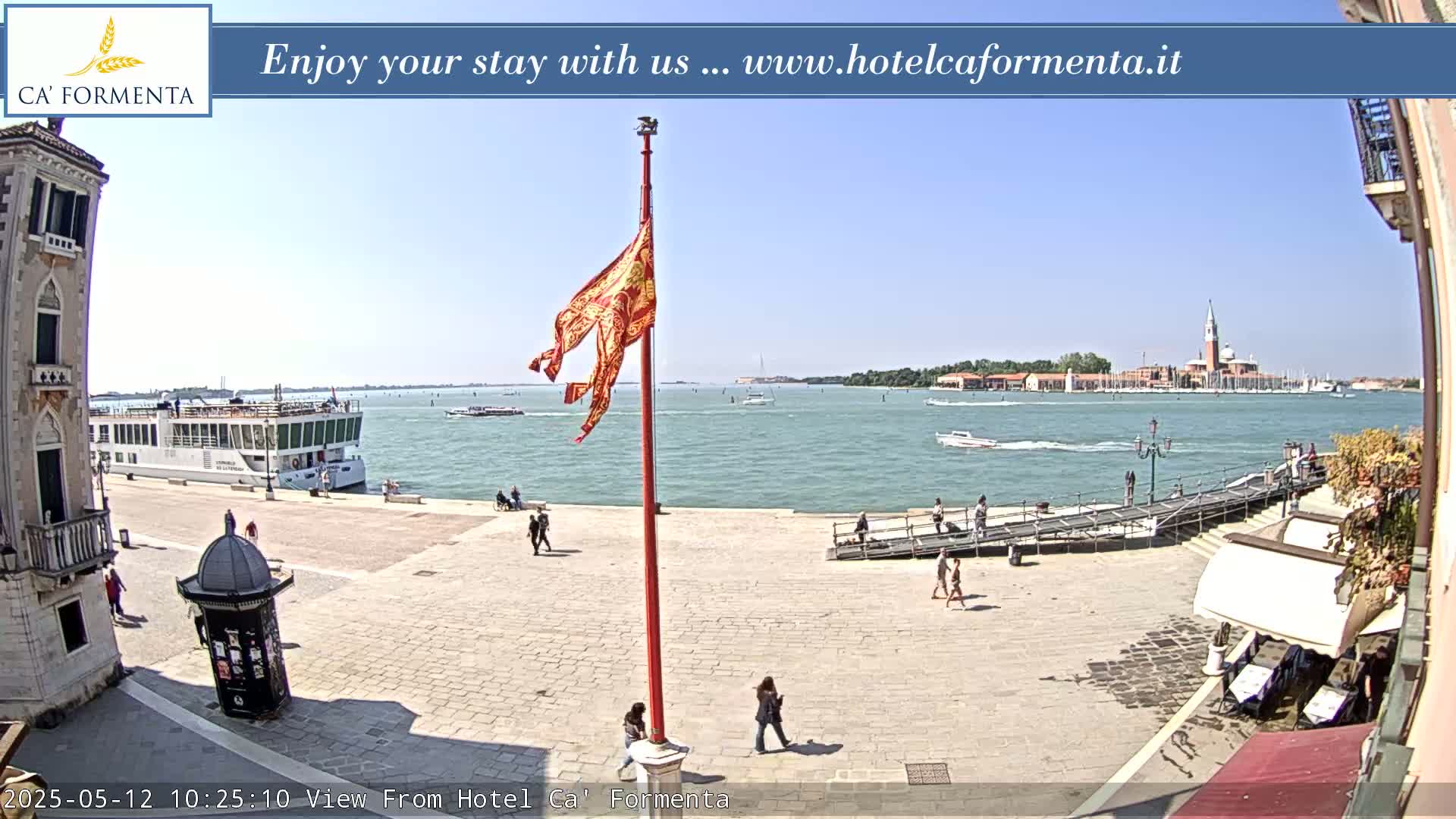 A sunny day in Venice shows a waterfront plaza with a large cruise ship and distant island, a flagpole with a red and gold flag, and a few people walking around.