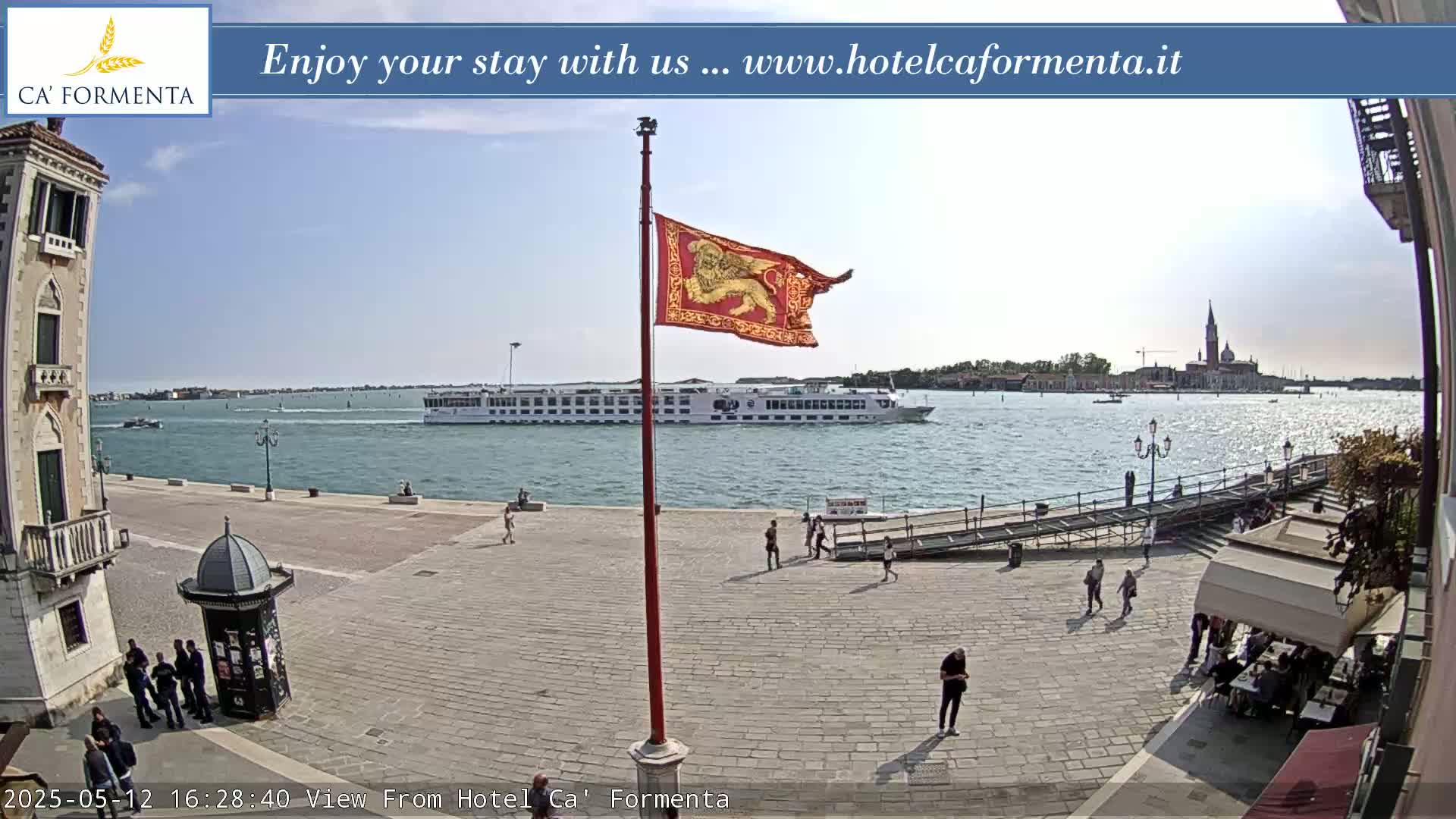 A sunny waterfront plaza shows a large cruise ship in the distance, a red flag with a lion emblem on a pole, and several people walking around.