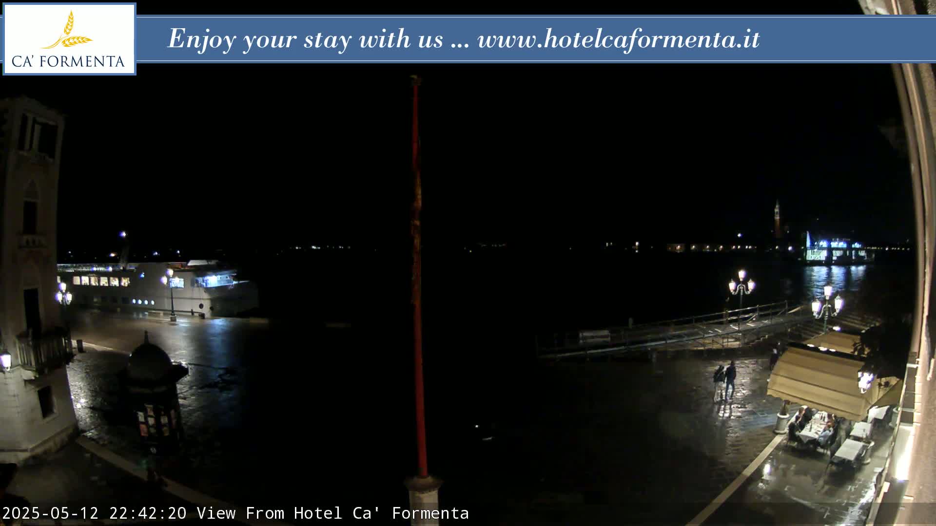 A nighttime view of a waterfront plaza shows a wet cobblestone surface, a docked boat, and a few people near outdoor restaurant seating, with distant lights across the water.