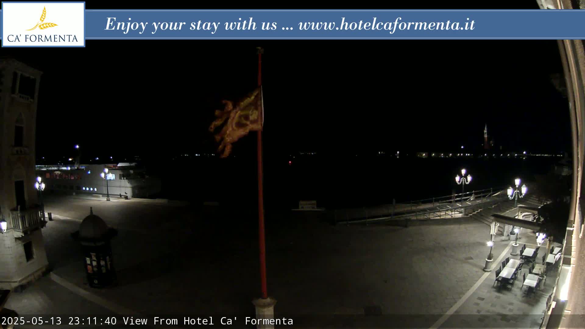 A nighttime view of a plaza with a flagpole, some outdoor seating, and a body of water with lights in the distance.